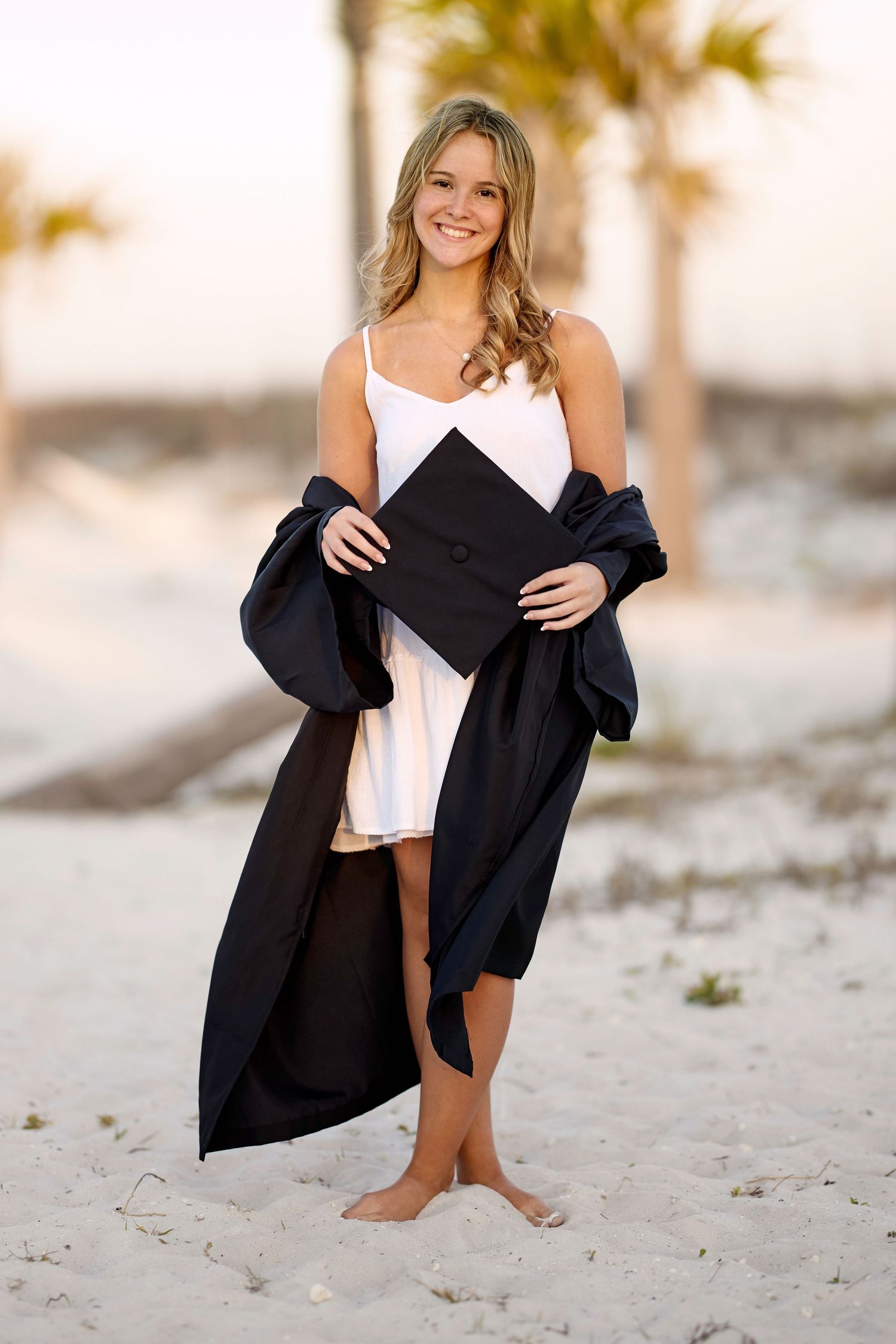 Smiling blonde graduate in white dress holding cap and black gown on a sandy beach with palm trees at sunset.