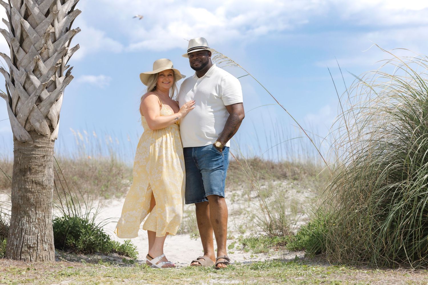 Couple posing near palm tree on sandy beach, woman in yellow jumpsuit and hat, man in white shirt and denim shorts.