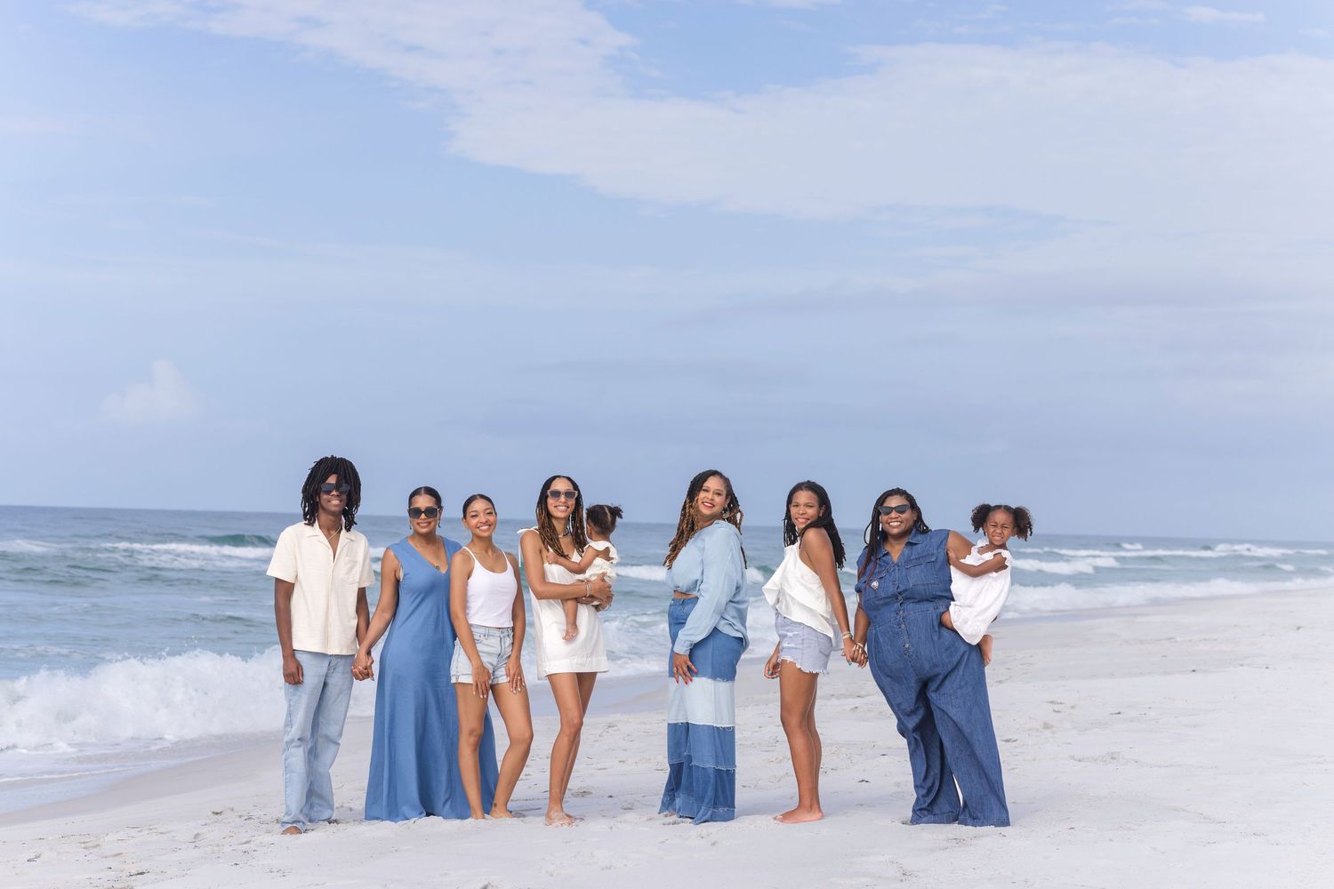 Group of eight people posing together on a white sand beach in blue and white outfits with ocean waves behind them.