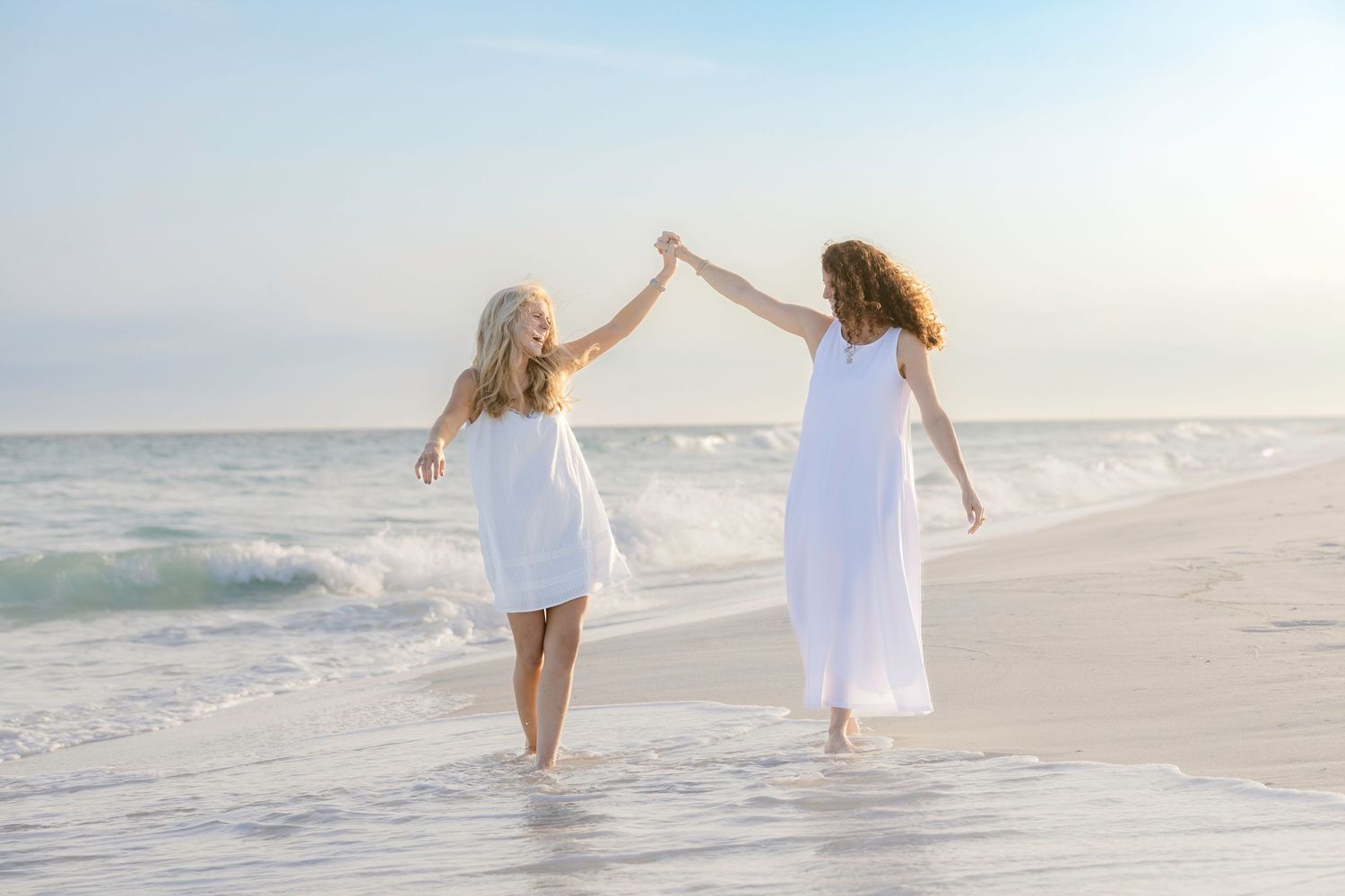 Two women in white dresses holding hands and dancing on a sunny beach with ocean waves behind them.