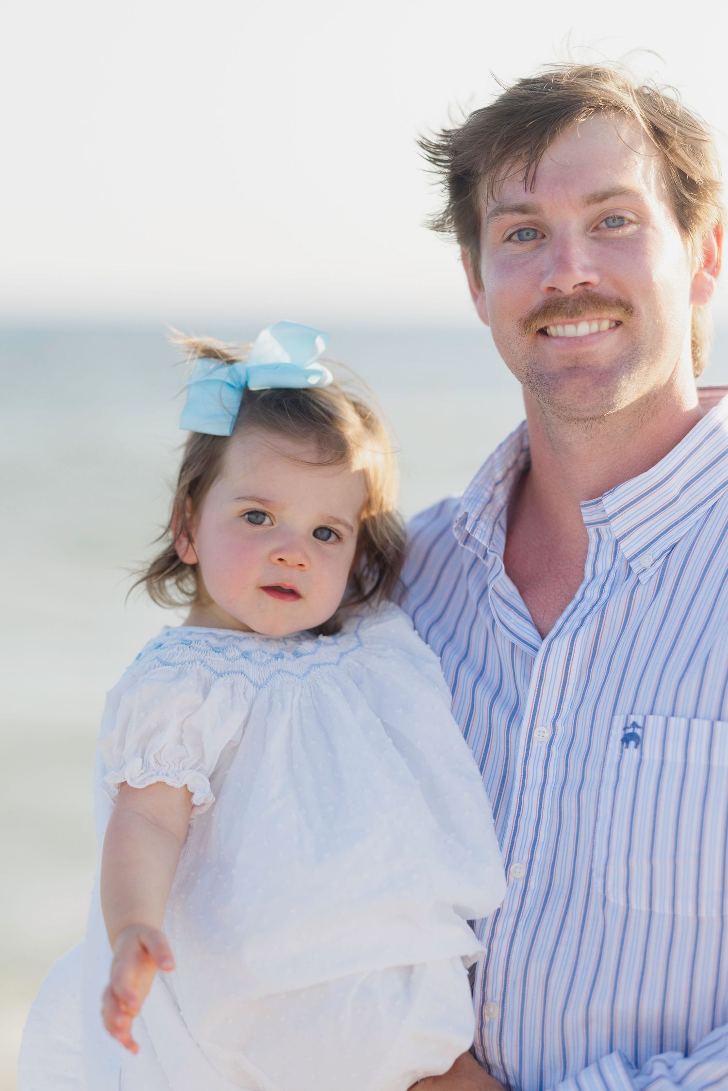 Father holding toddler daughter in white dress with blue bow at beach, smiling family portrait outdoors.