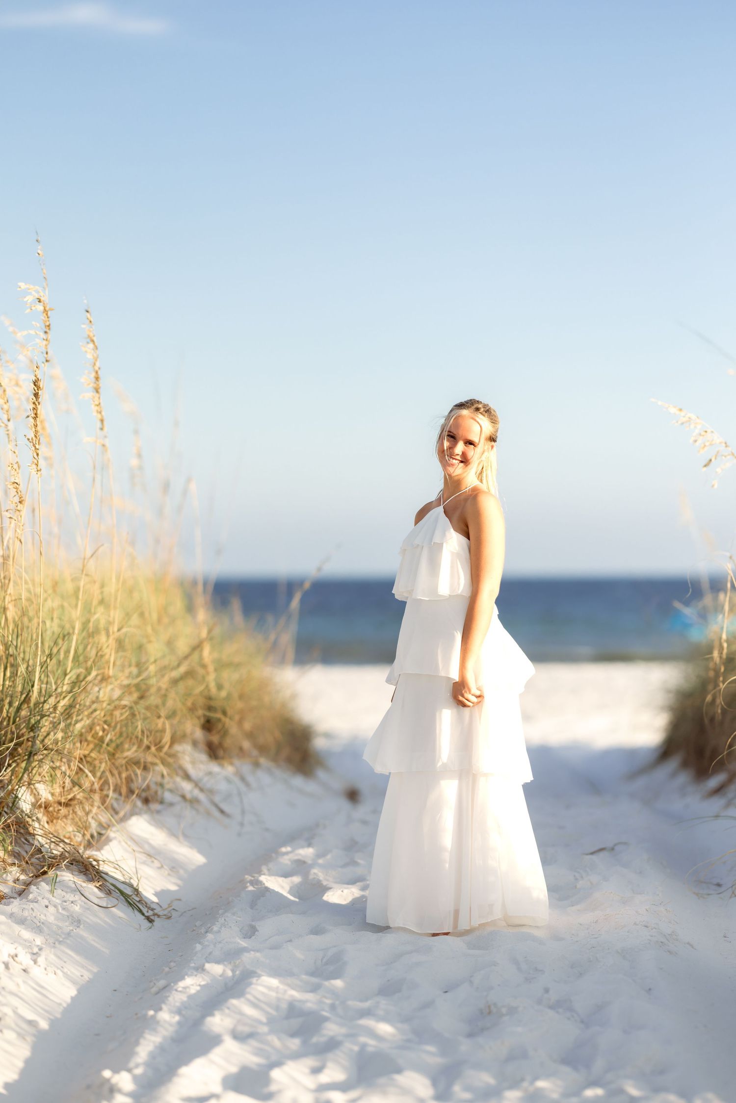 Bride in white tiered gown standing on sandy beach path with sea oats, ocean in background.