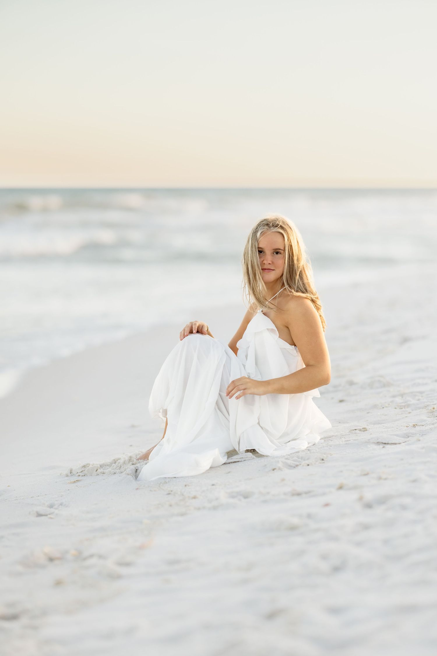 A blonde woman in a white strapless dress sits gracefully on a sandy beach with ocean waves in the background.