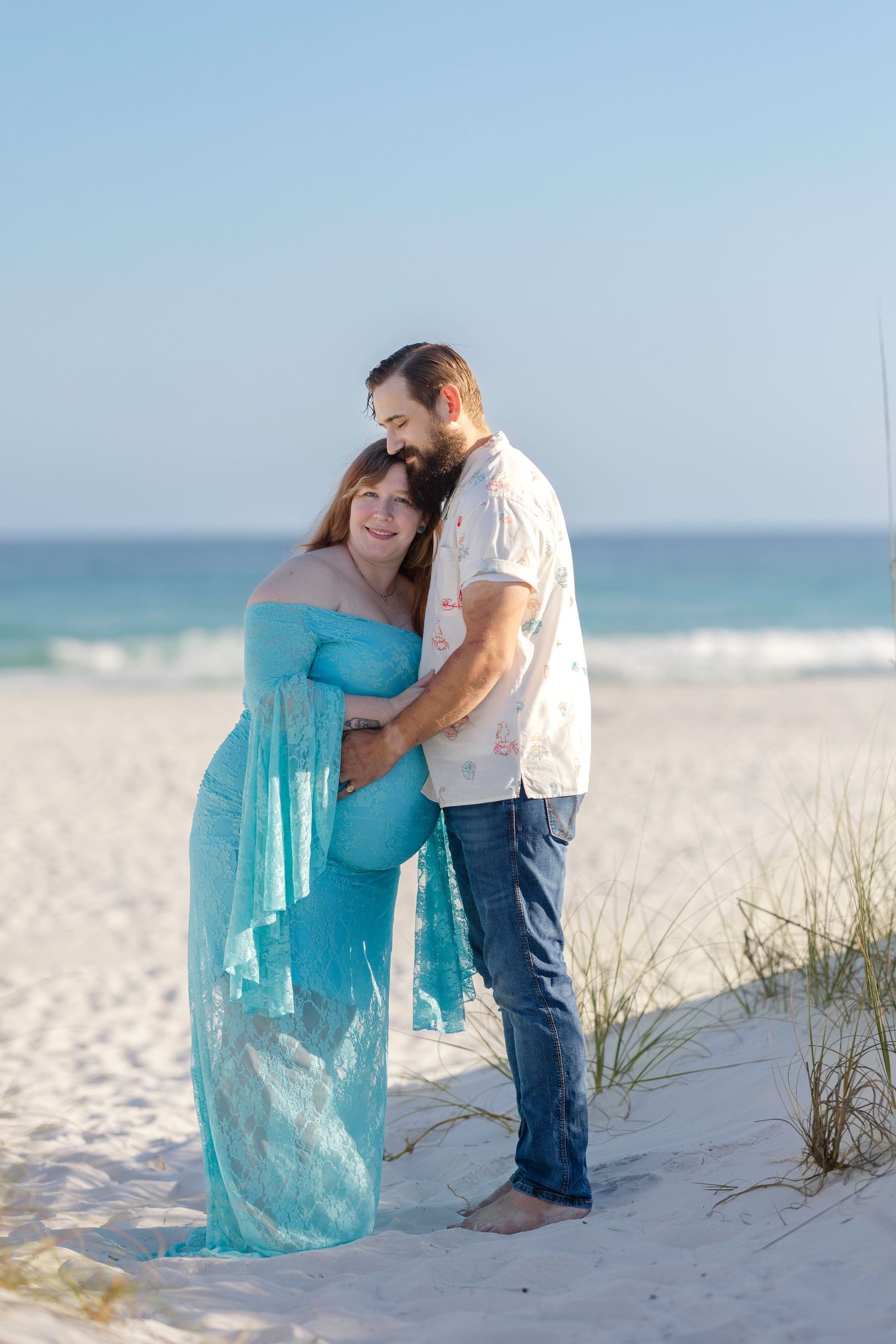 Pregnant woman in teal lace gown embraced by bearded man on white sand beach with ocean backdrop.