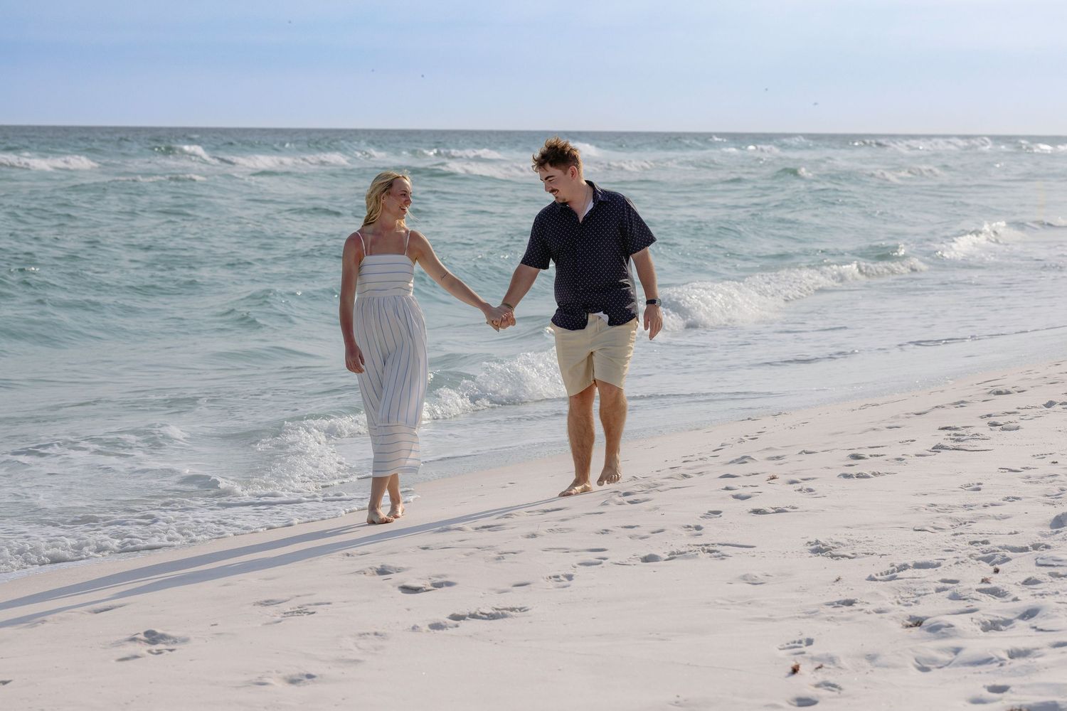 Couple holding hands walking barefoot on a white sandy beach with turquoise ocean waves in the background.