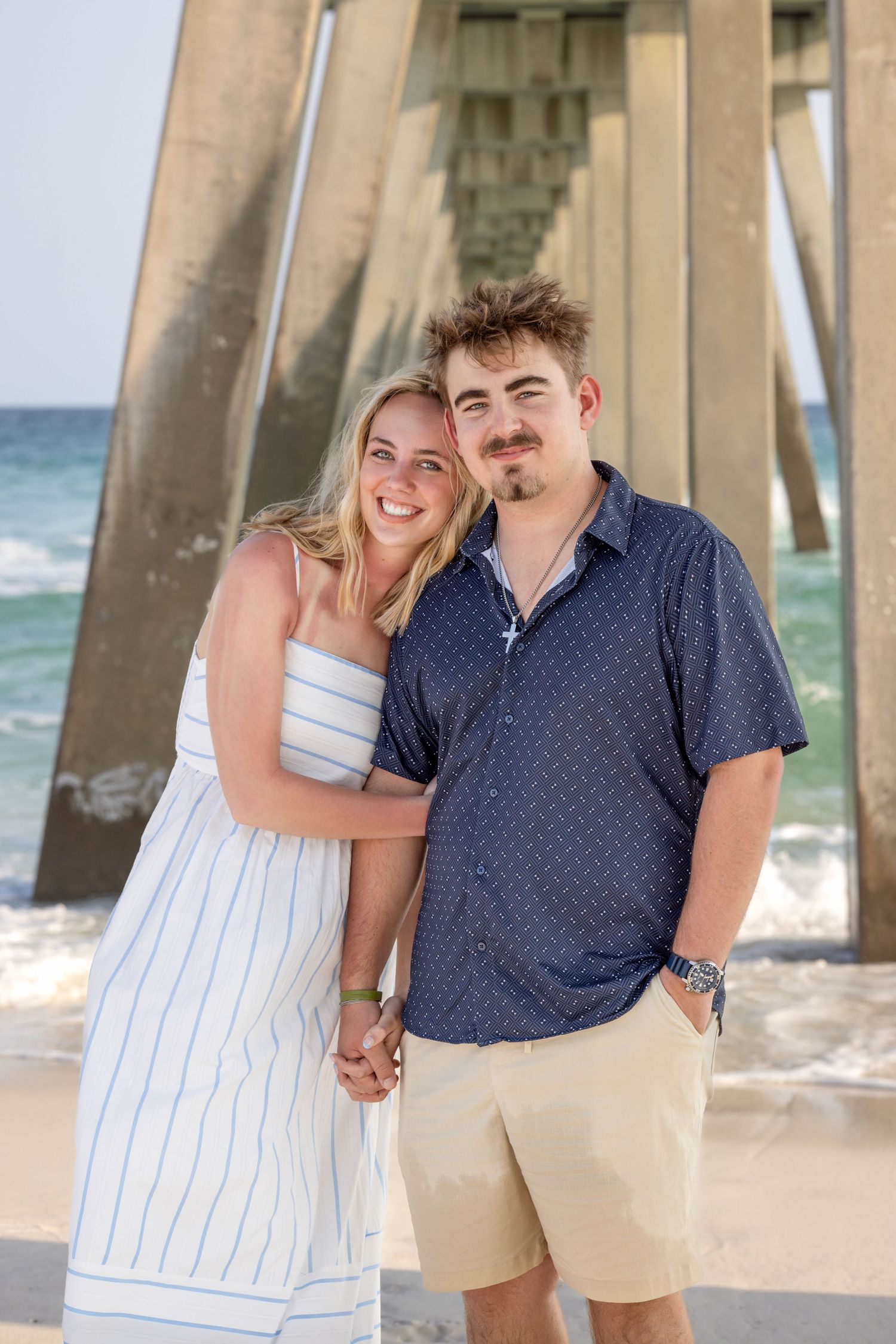 A smiling couple poses on a beach near a pier, woman in white dress, man in navy shirt and khaki shorts.