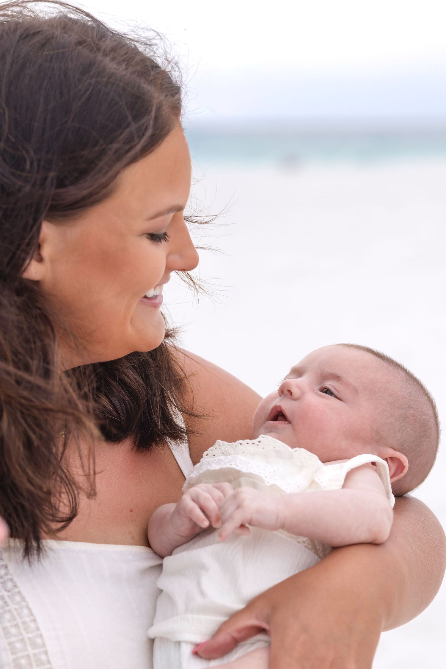 Smiling mother holding her newborn baby on a bright beach, both dressed in white.