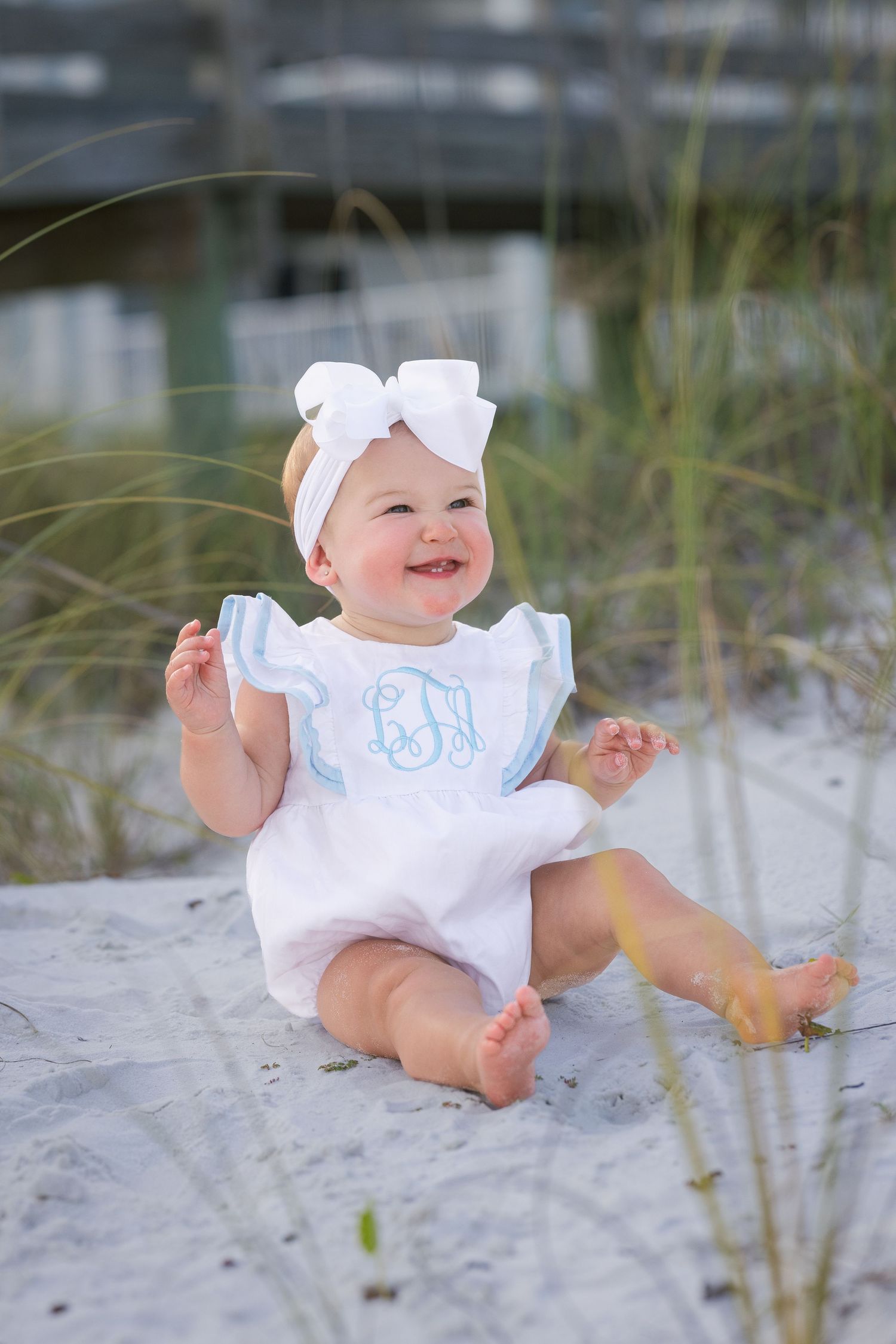 Happy baby girl in white monogrammed romper with bow headband sitting on sandy beach near dunes.