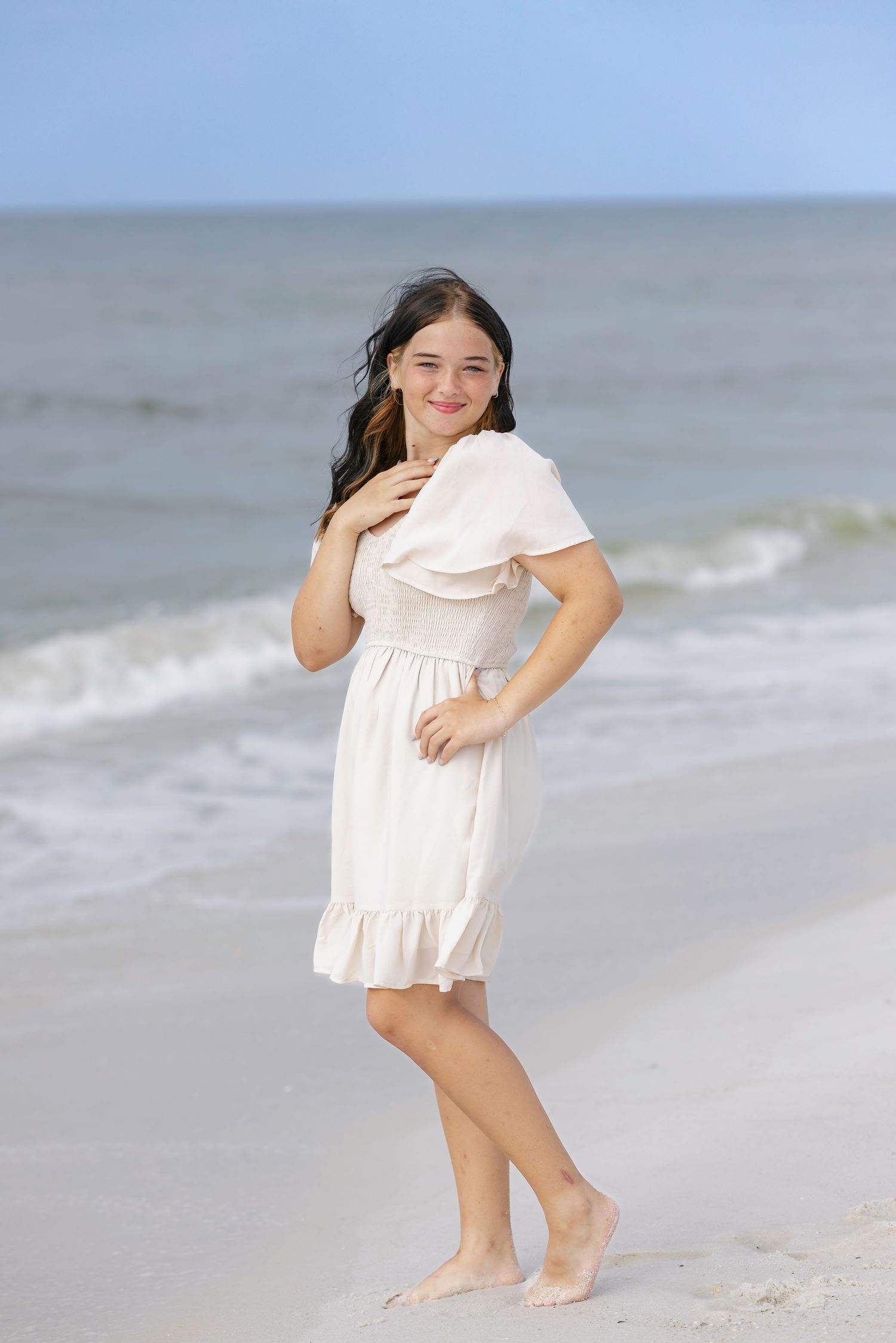 Young girl in white dress posing barefoot on sandy beach with ocean waves in background.