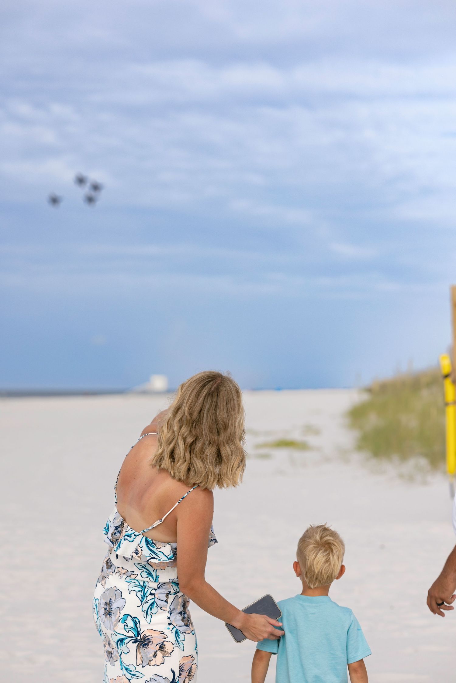 A woman and young boy watch military jets fly over a white sandy beach on a cloudy day.
