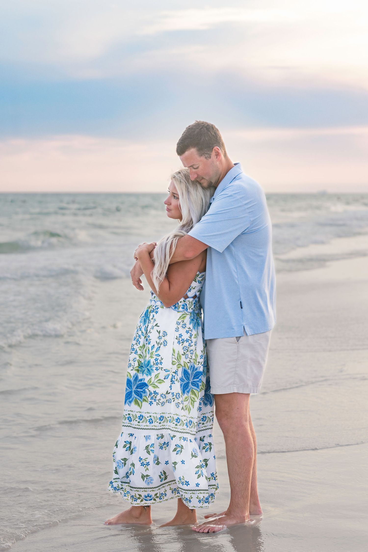 Couple embracing on a beach at sunset, woman in floral blue dress, man in light blue shirt and white shorts.