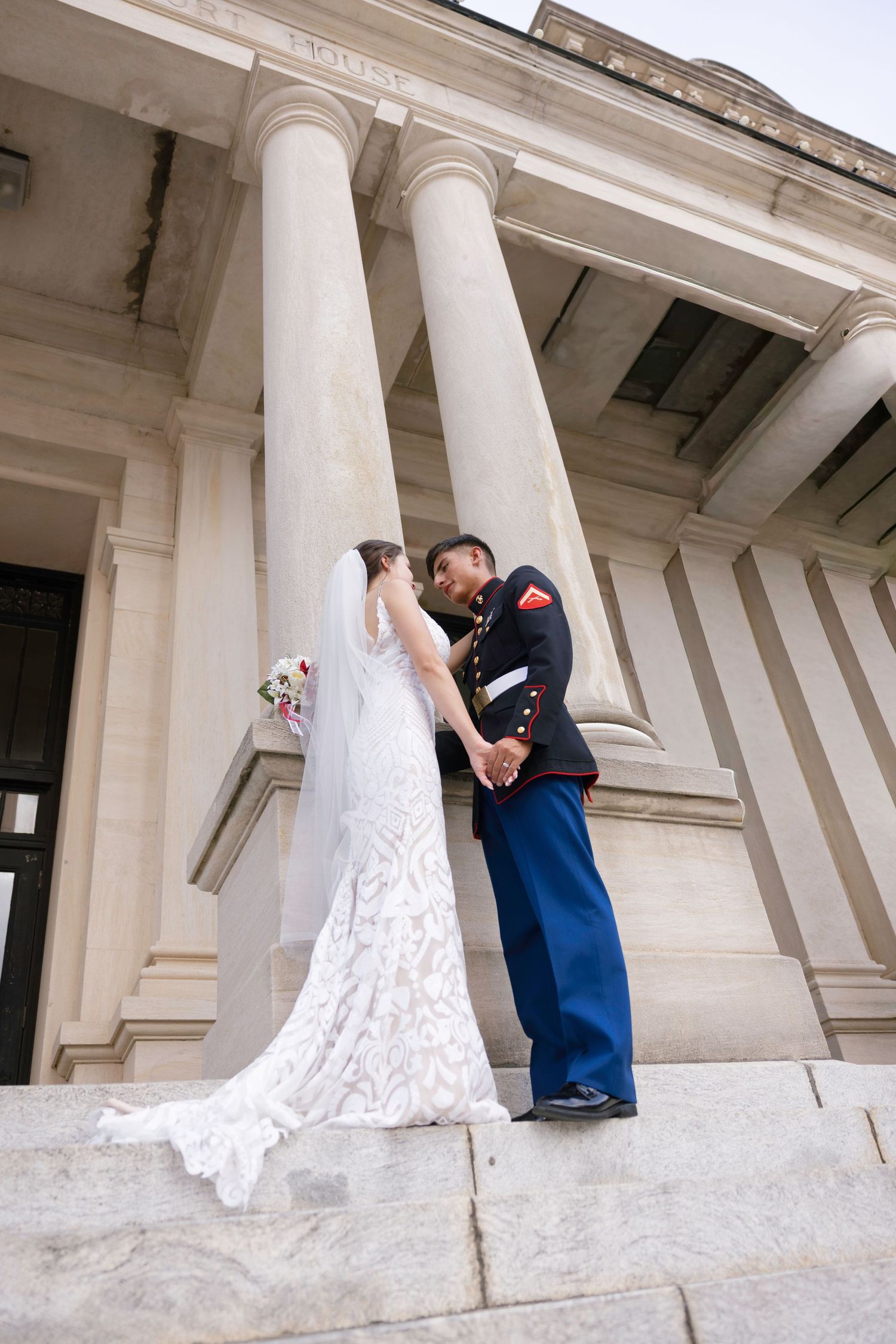 Marine in dress uniform and bride in lace gown share a romantic moment on grand building steps with tall columns.
