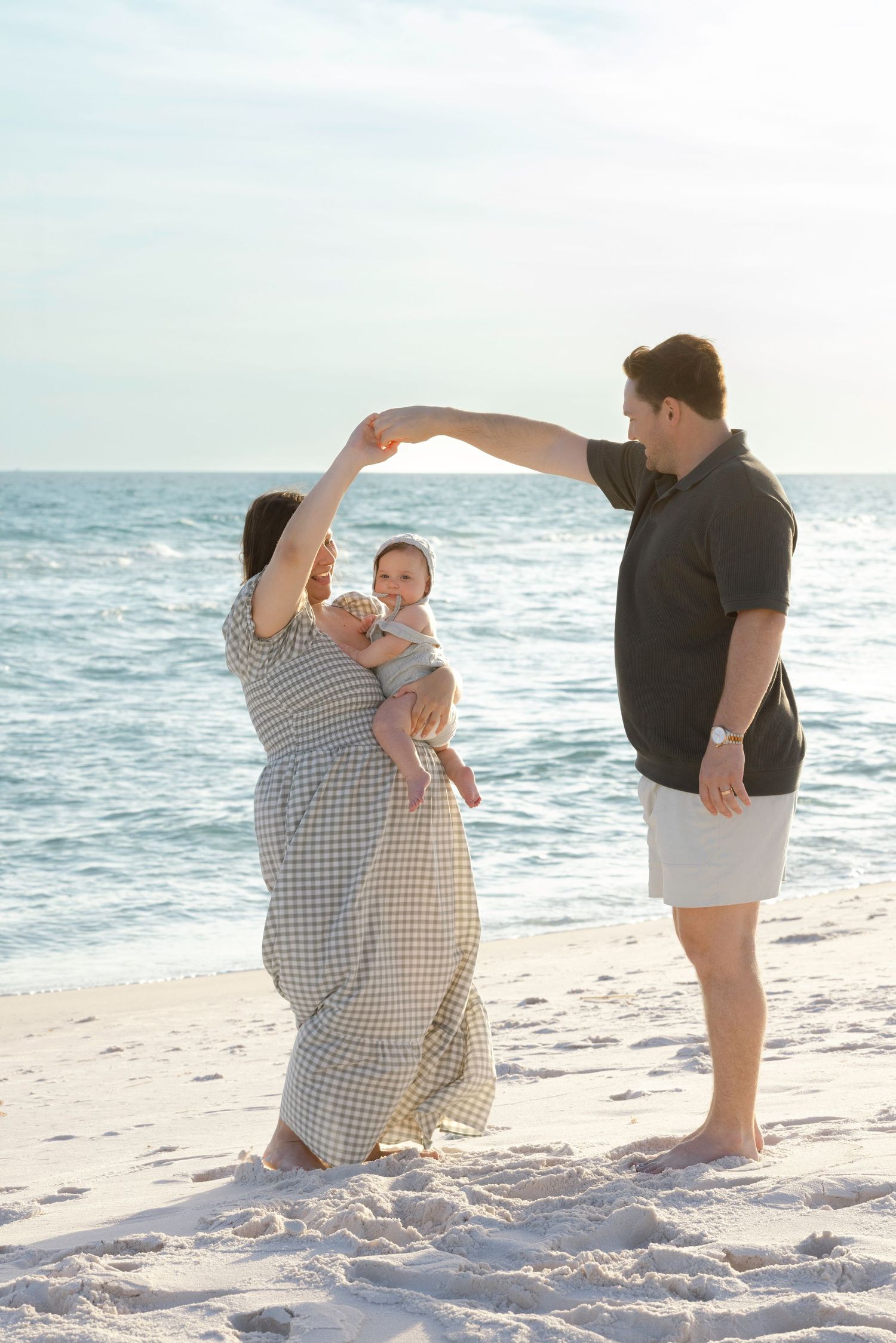 A couple dances on a sandy beach while holding their baby, with calm ocean waves in the background.