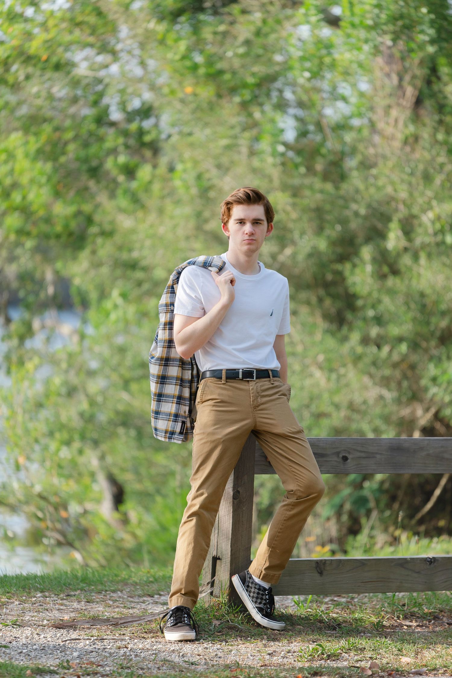 Young man in white tee and tan chinos leaning on wooden fence outdoors, holding plaid jacket over shoulder.