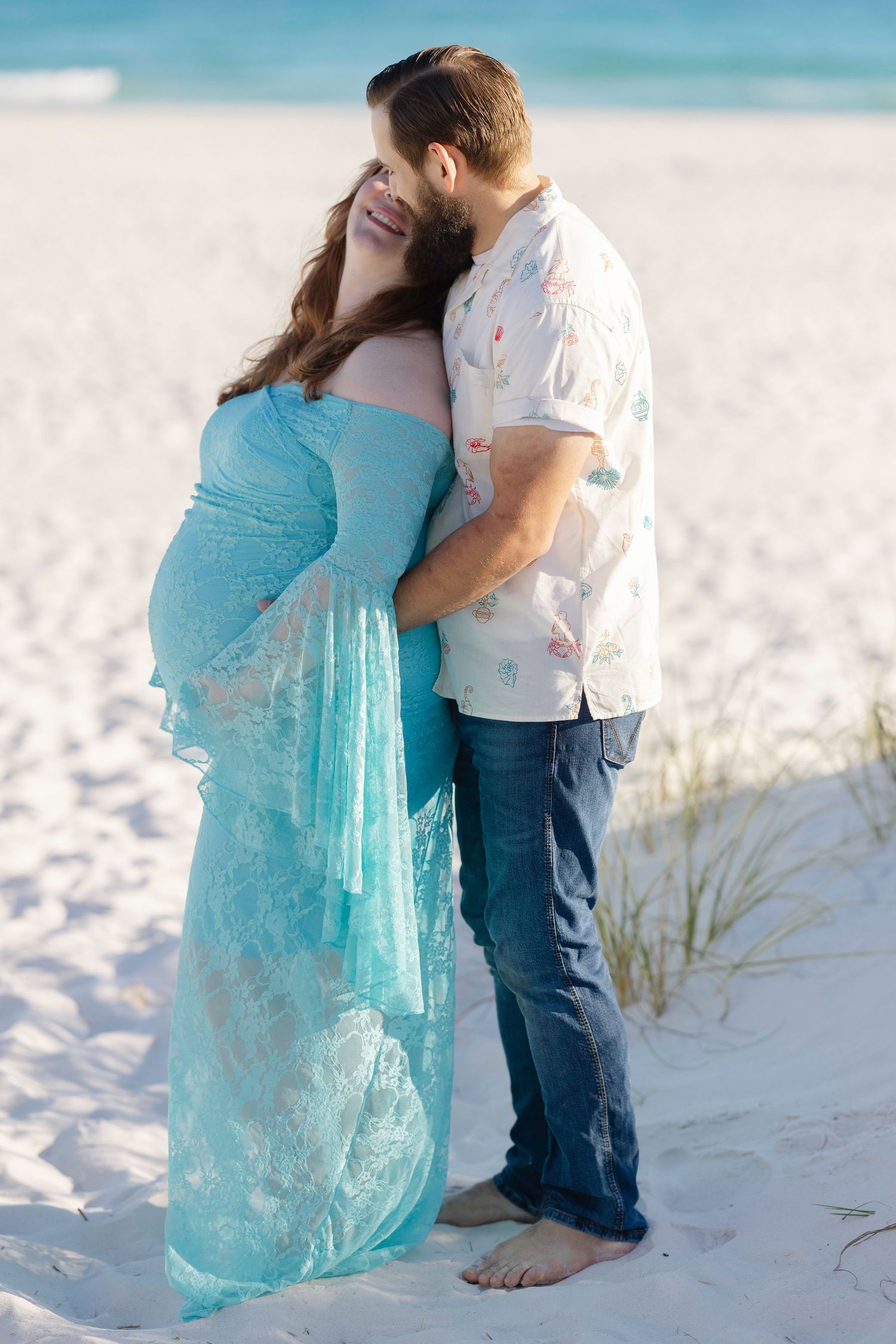 Pregnant woman in teal lace gown embraced by bearded man on white sand beach for maternity photo.