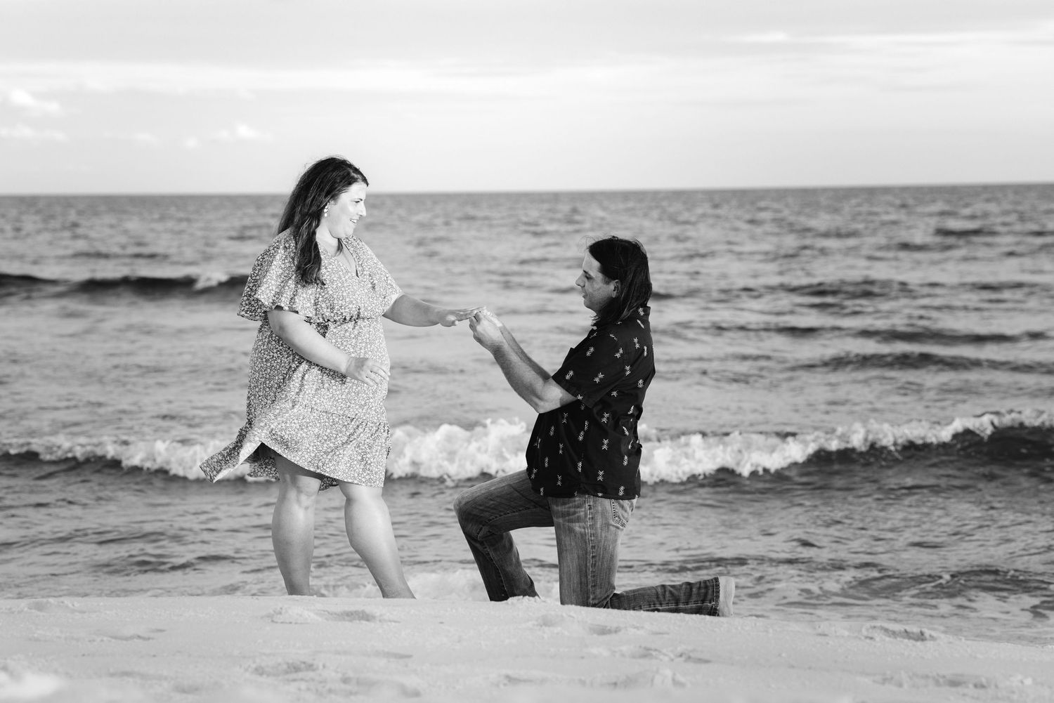 Black and white beach proposal photo of man on one knee holding woman's hand by ocean waves.