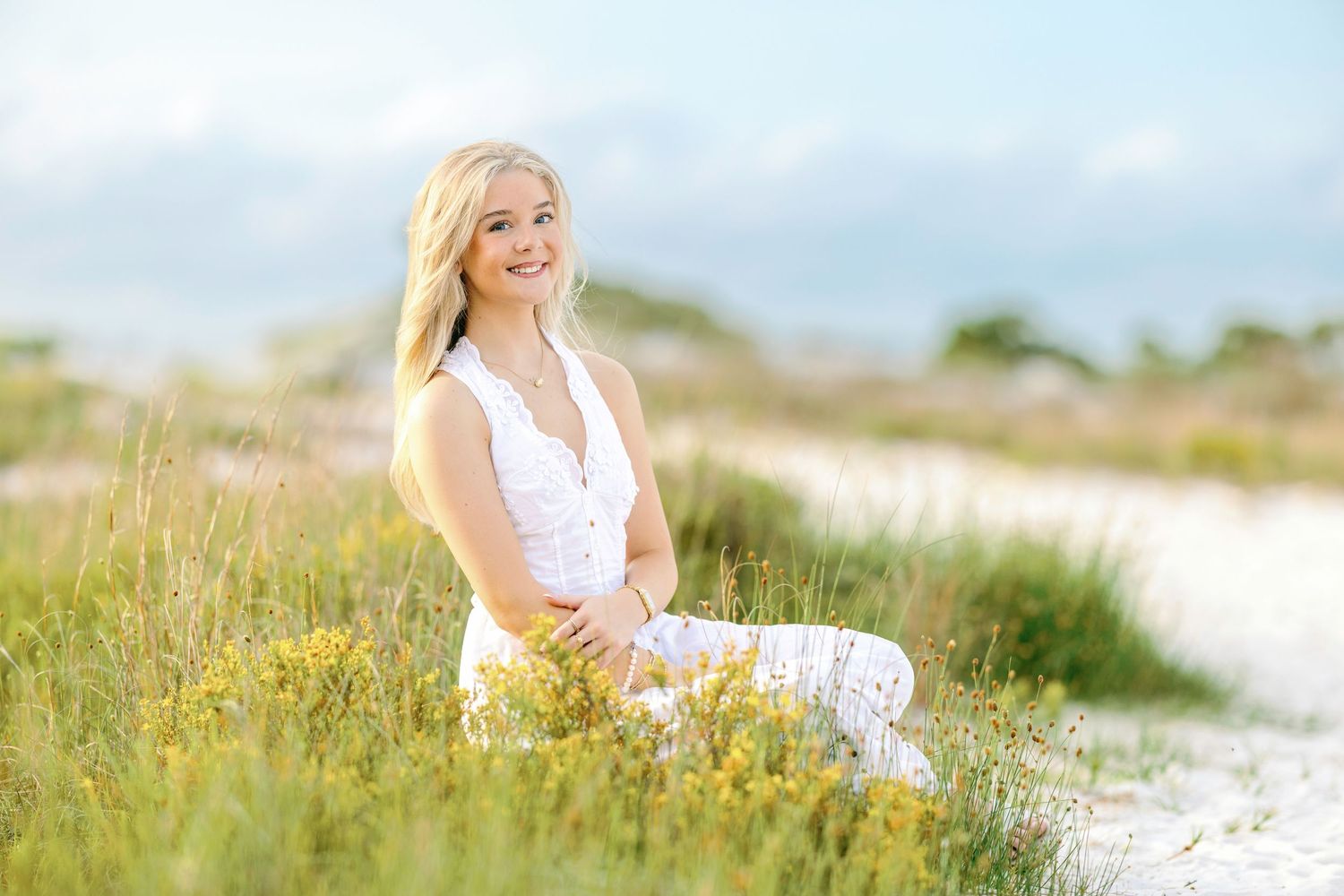 Smiling blonde woman in white outfit sitting in green meadow with wildflowers on a sunny day.