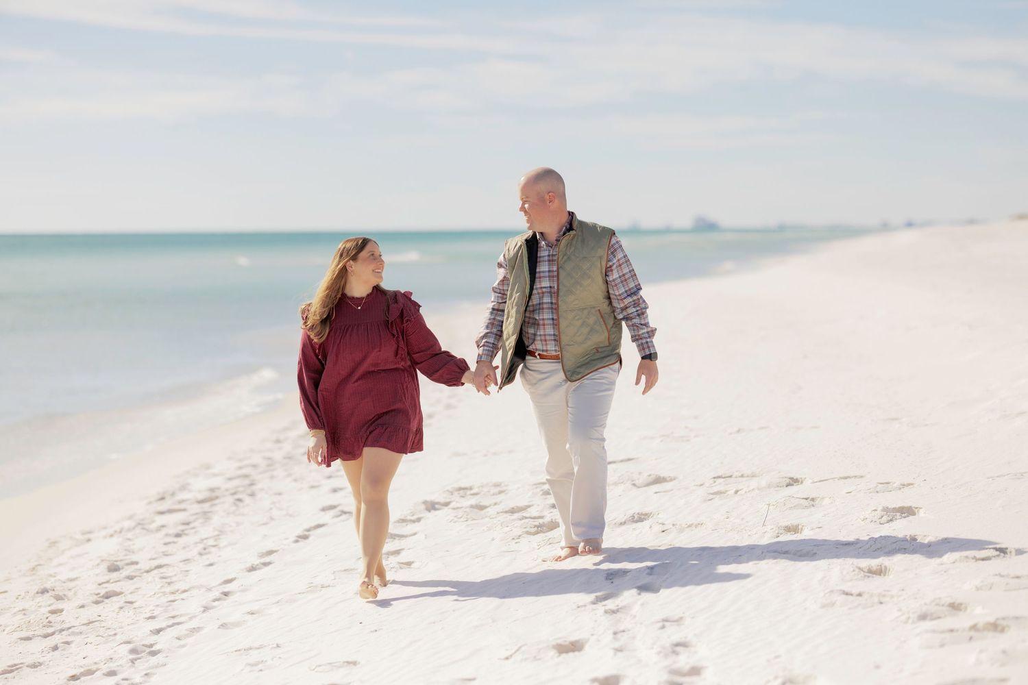 Couple holding hands walking on white sandy beach near turquoise ocean water on a sunny day.