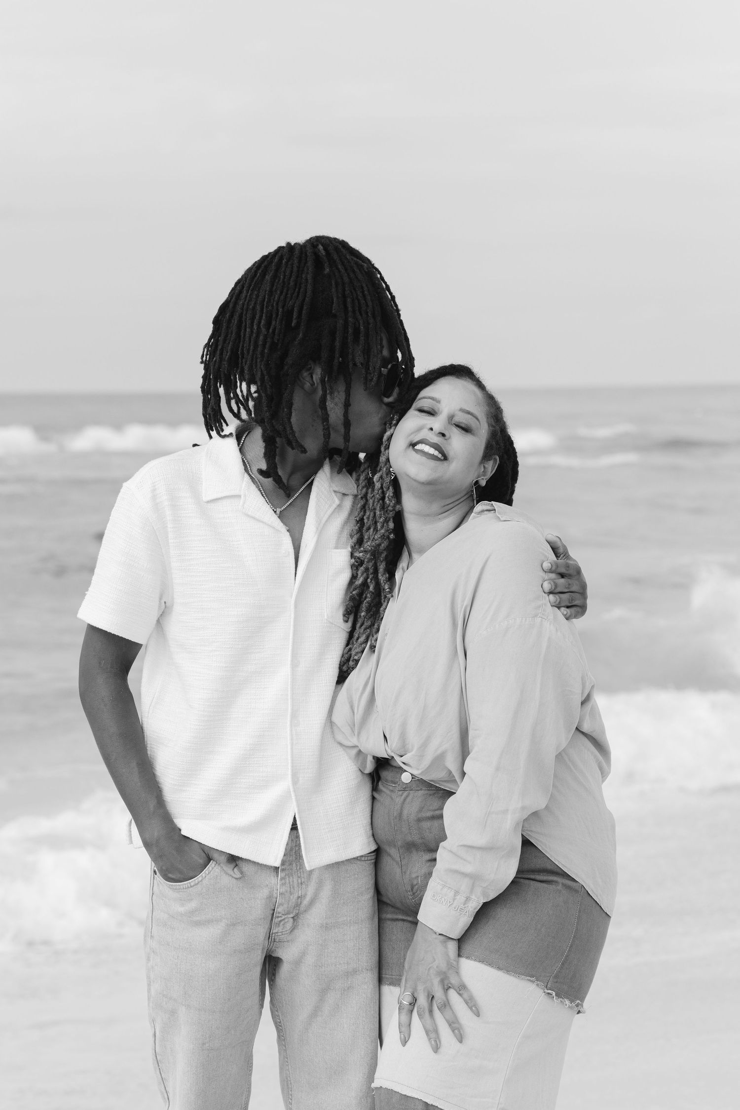 Black and white photo of a happy interracial couple embracing on a beach with ocean waves in the background.