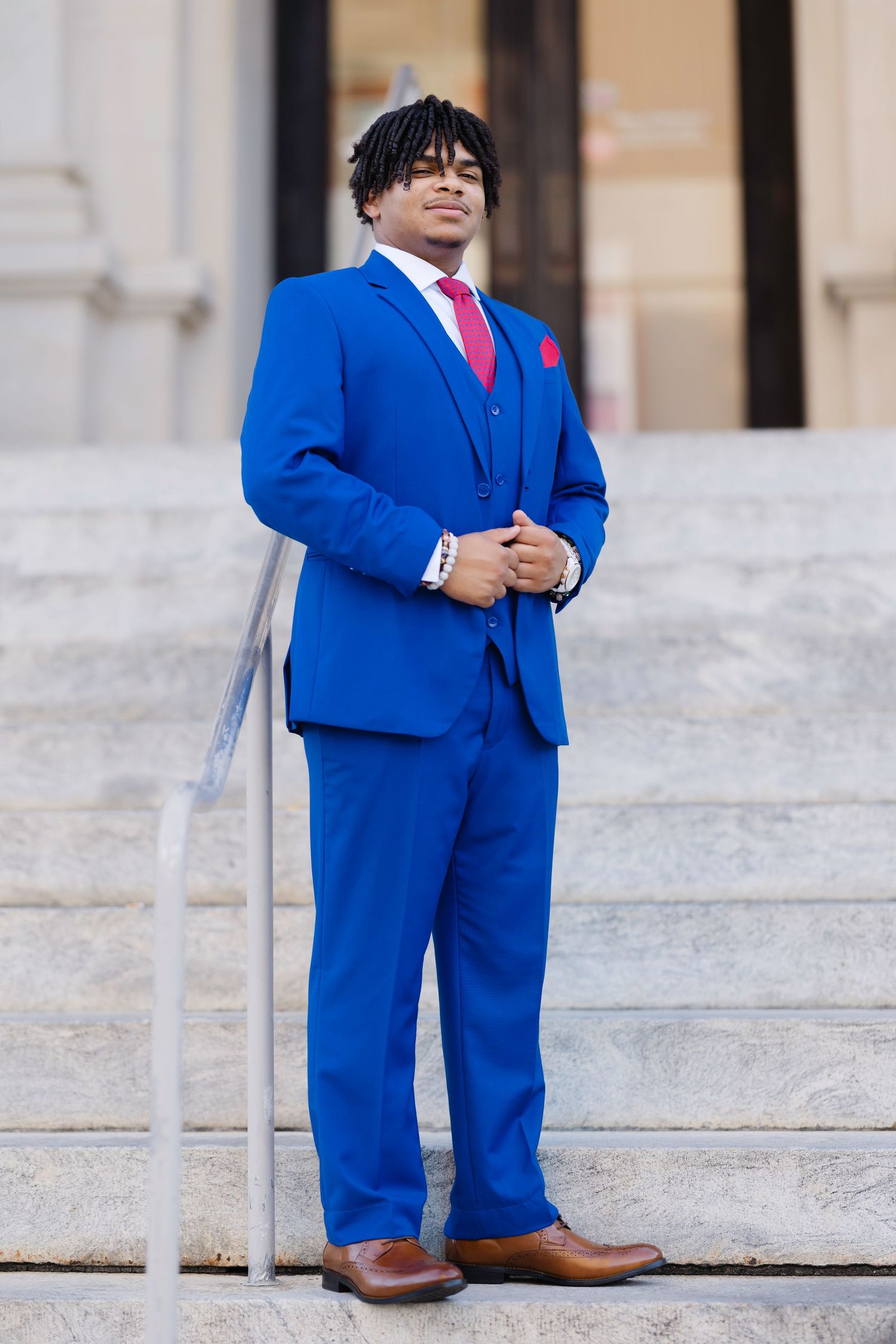Young man in a bold royal blue three-piece suit with pink tie and pocket square, standing on marble steps.