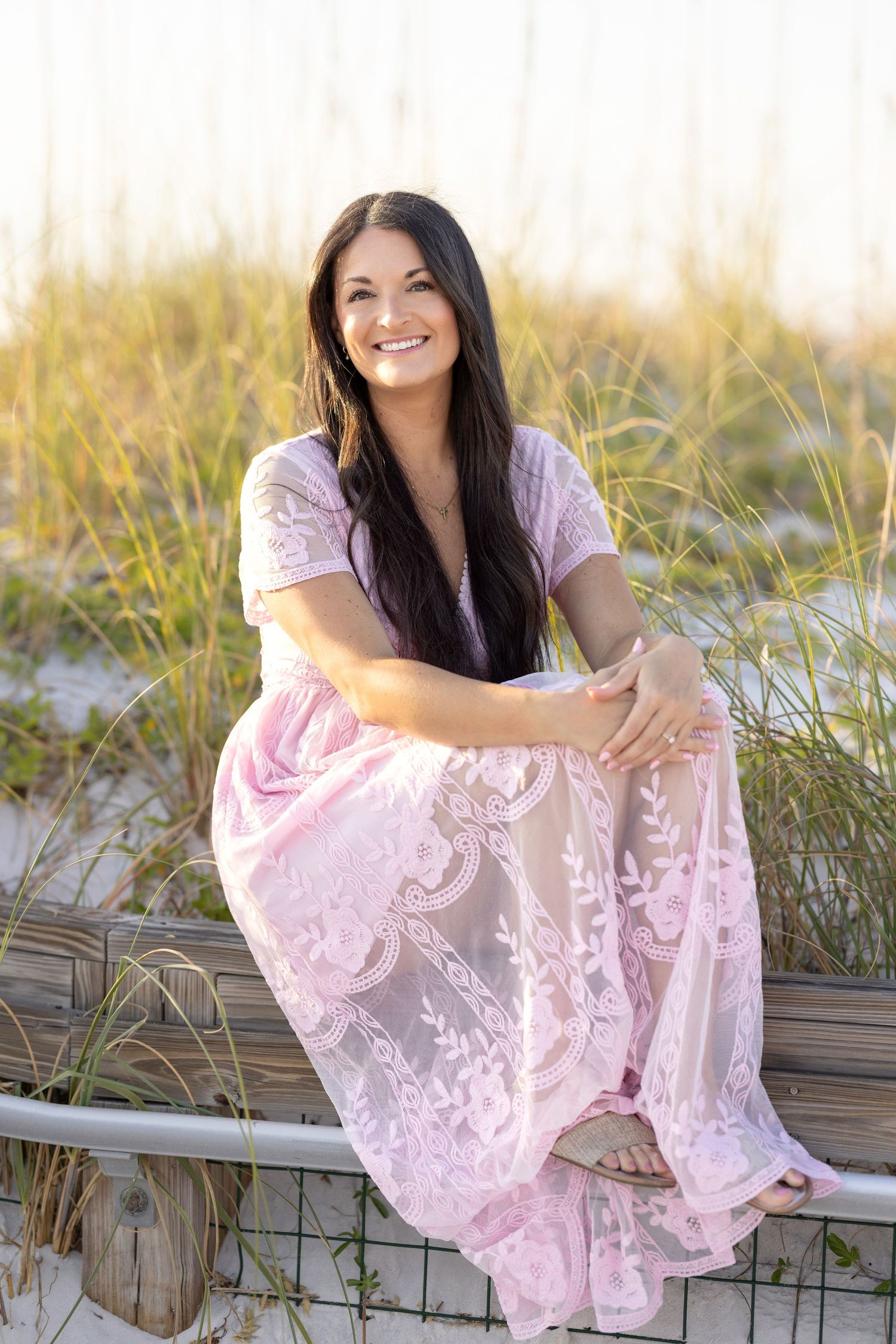 Woman in pink lace dress sitting on beach fence smiling with sand dunes and sea grass background.