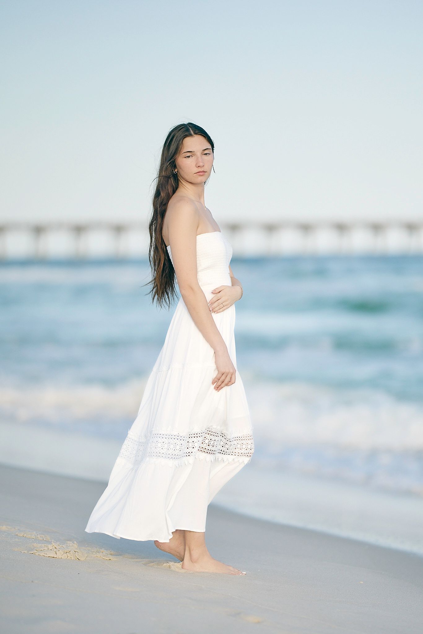 Person in flowy white dress walking barefoot along a serene beach with ocean waves in soft blue tones.