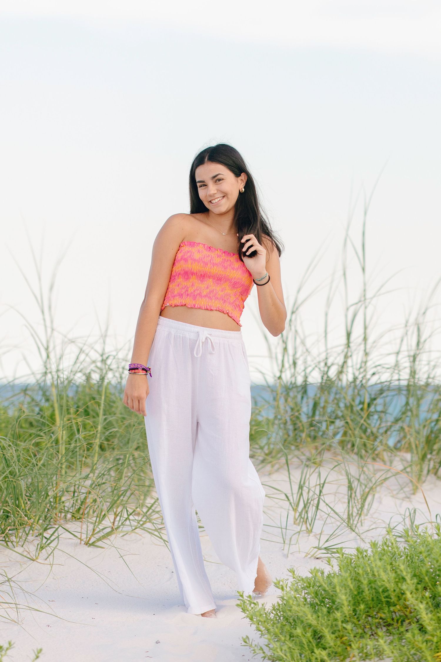 Woman in coral top and white pants poses gracefully on sandy beach with sea grass and ocean in background.
