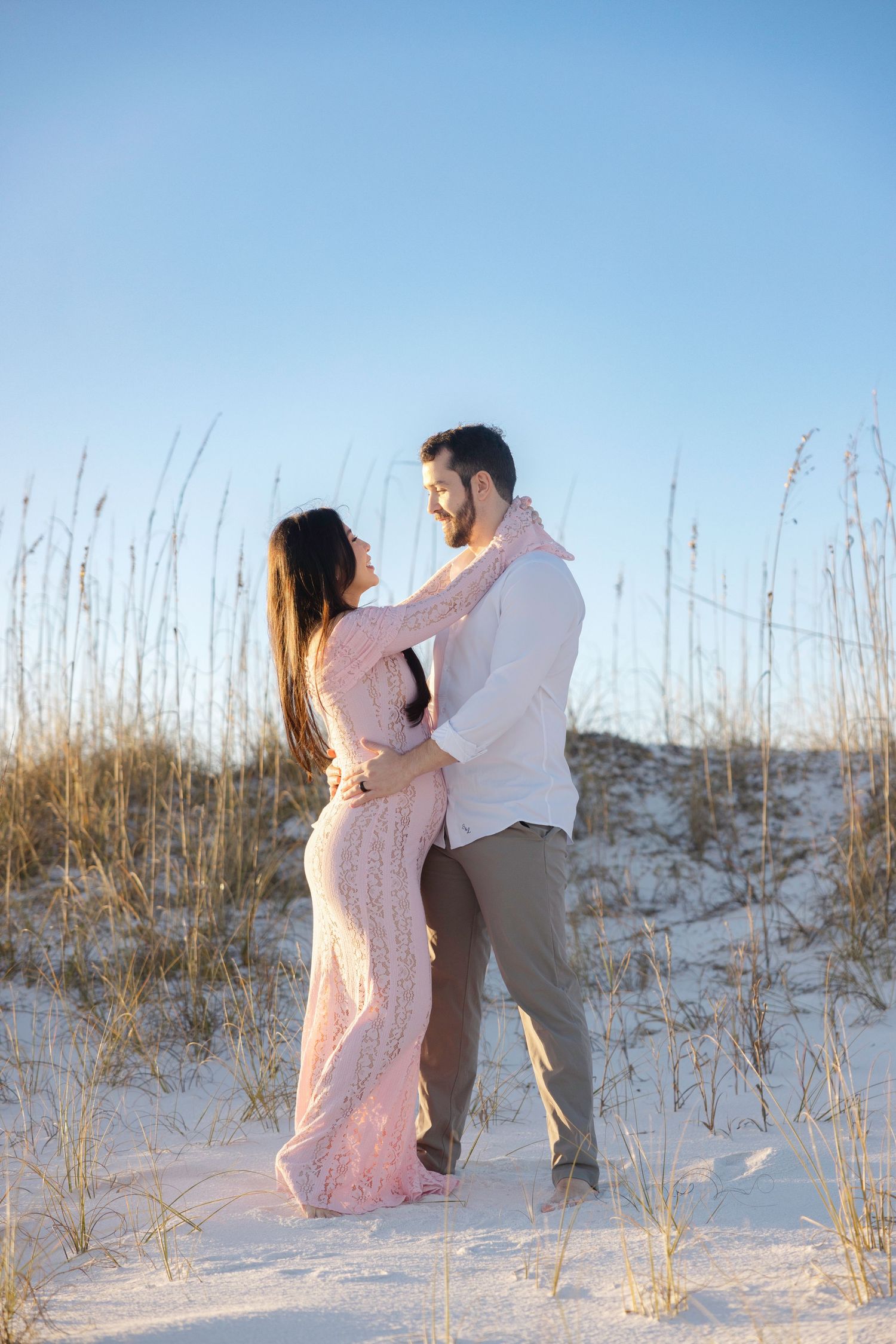 A romantic couple embraces on white sand dunes with tall beach grass at sunset.