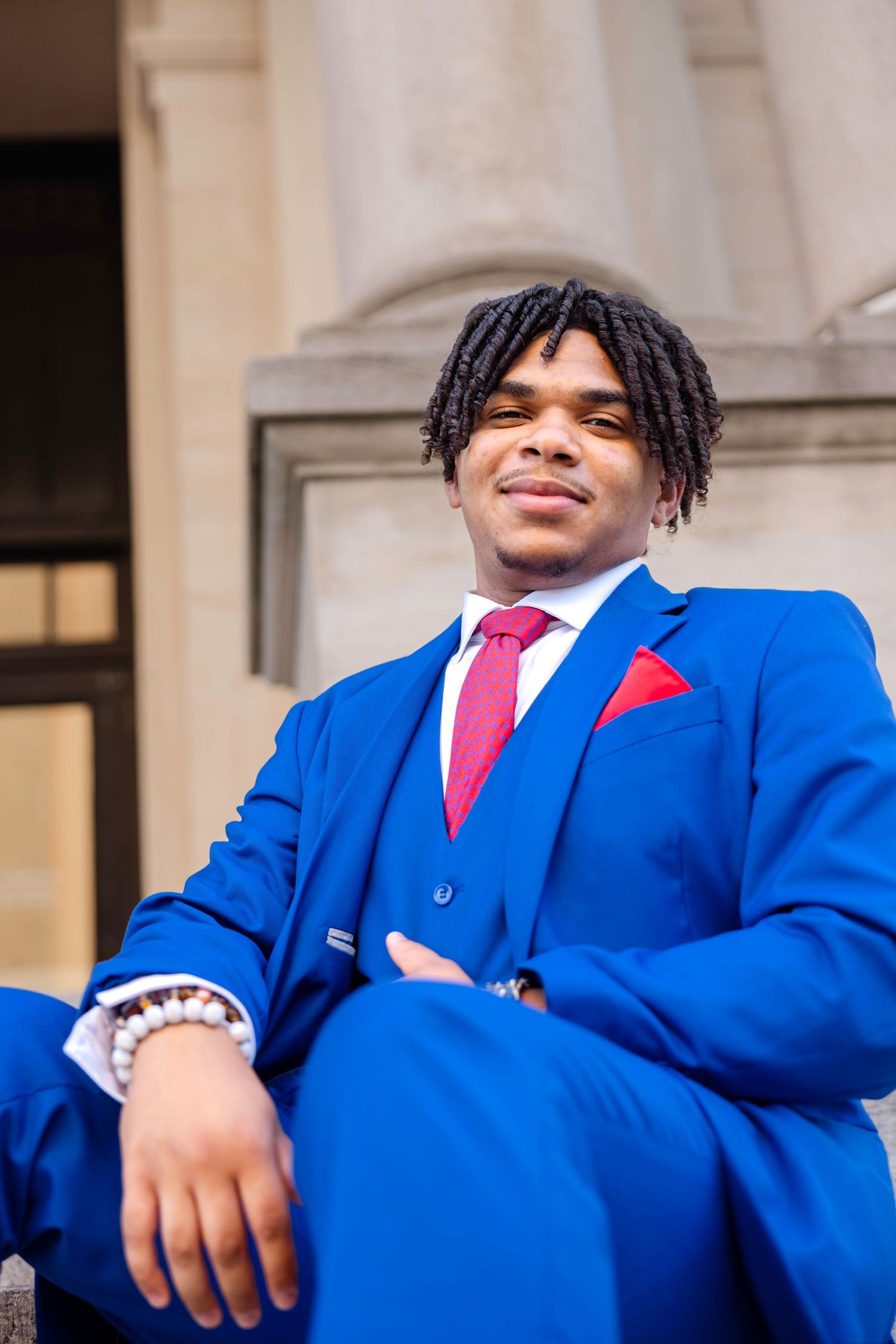 Confident young Black man in vibrant blue three-piece suit with red tie and pocket square relaxes on stone steps.