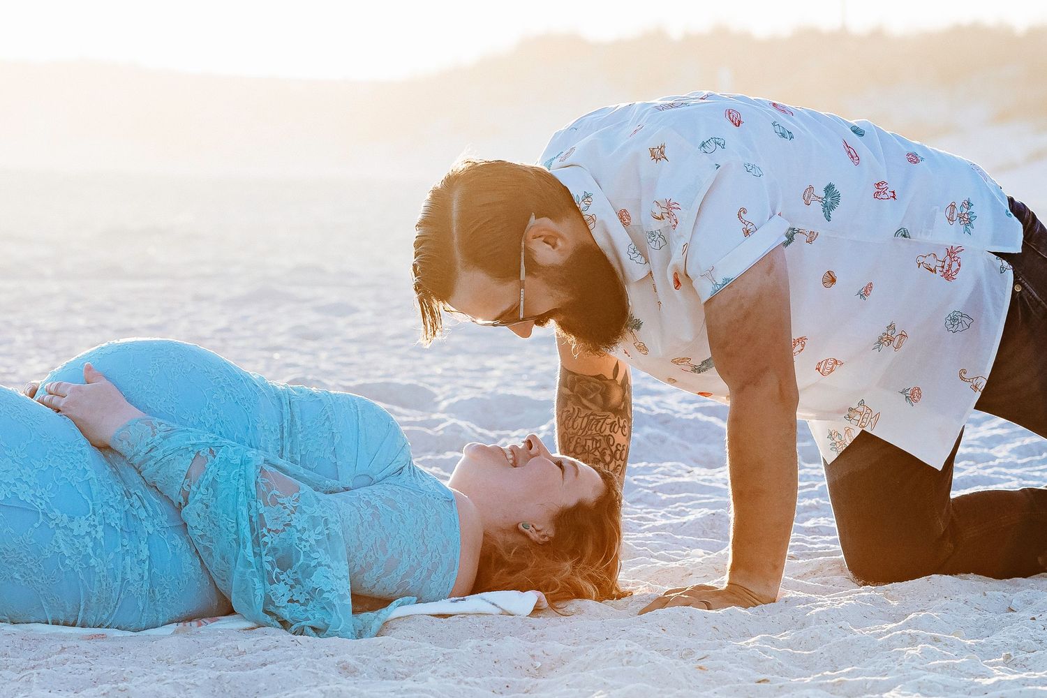 Couple sharing a tender moment on a sandy beach at golden hour, woman lying down smiling up at man.
