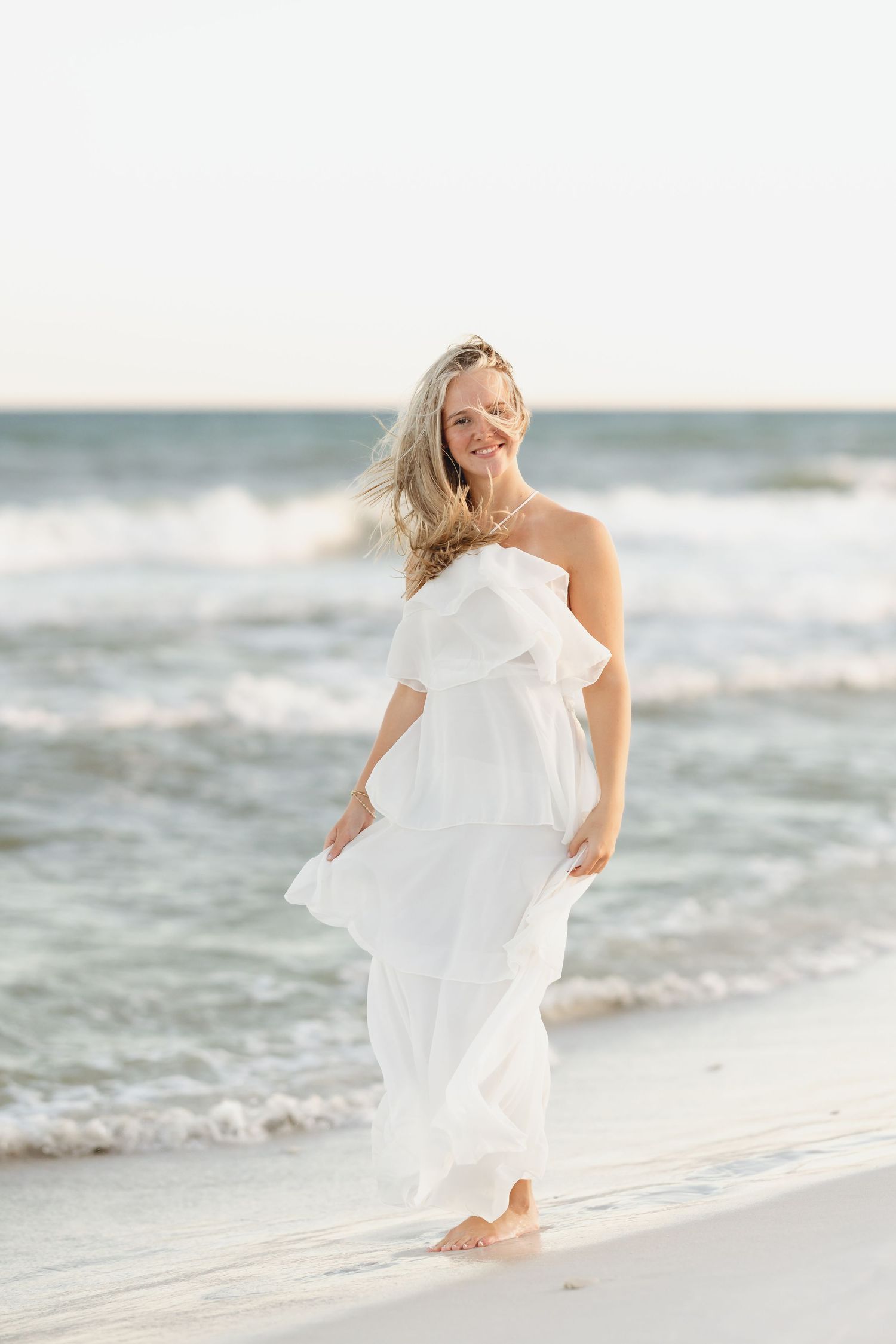 A smiling blonde woman in a white strapless ruffled dress walks barefoot on a sandy beach with ocean waves behind her.