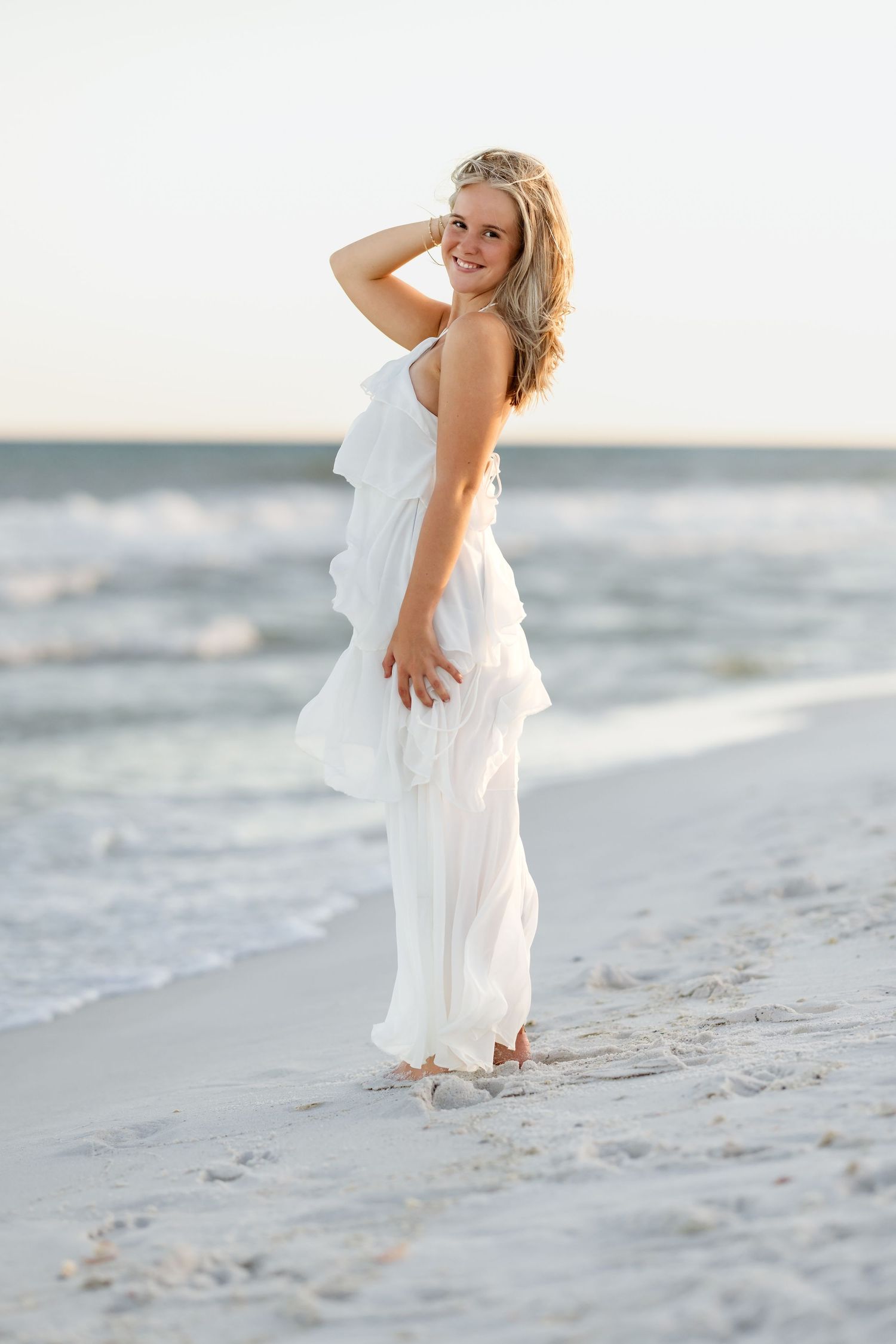 Blonde woman in white ruffled maxi dress poses on beach shoreline at sunset, hand in hair, smiling over shoulder.