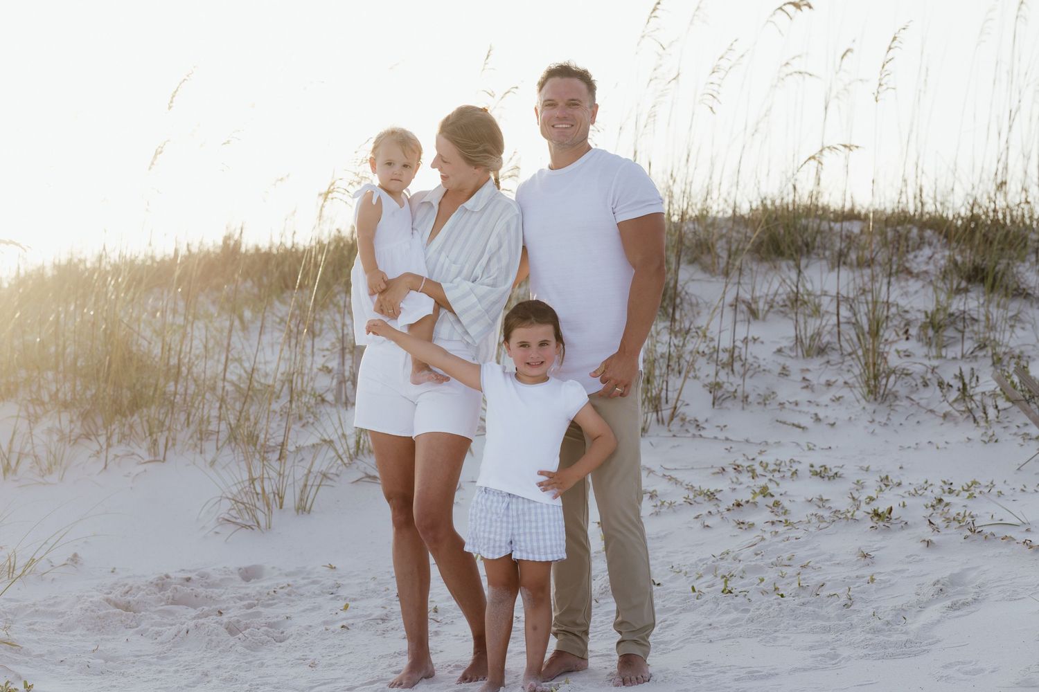 Family of four in white outfits posing on sandy beach dunes with sea oats in warm golden sunlight.