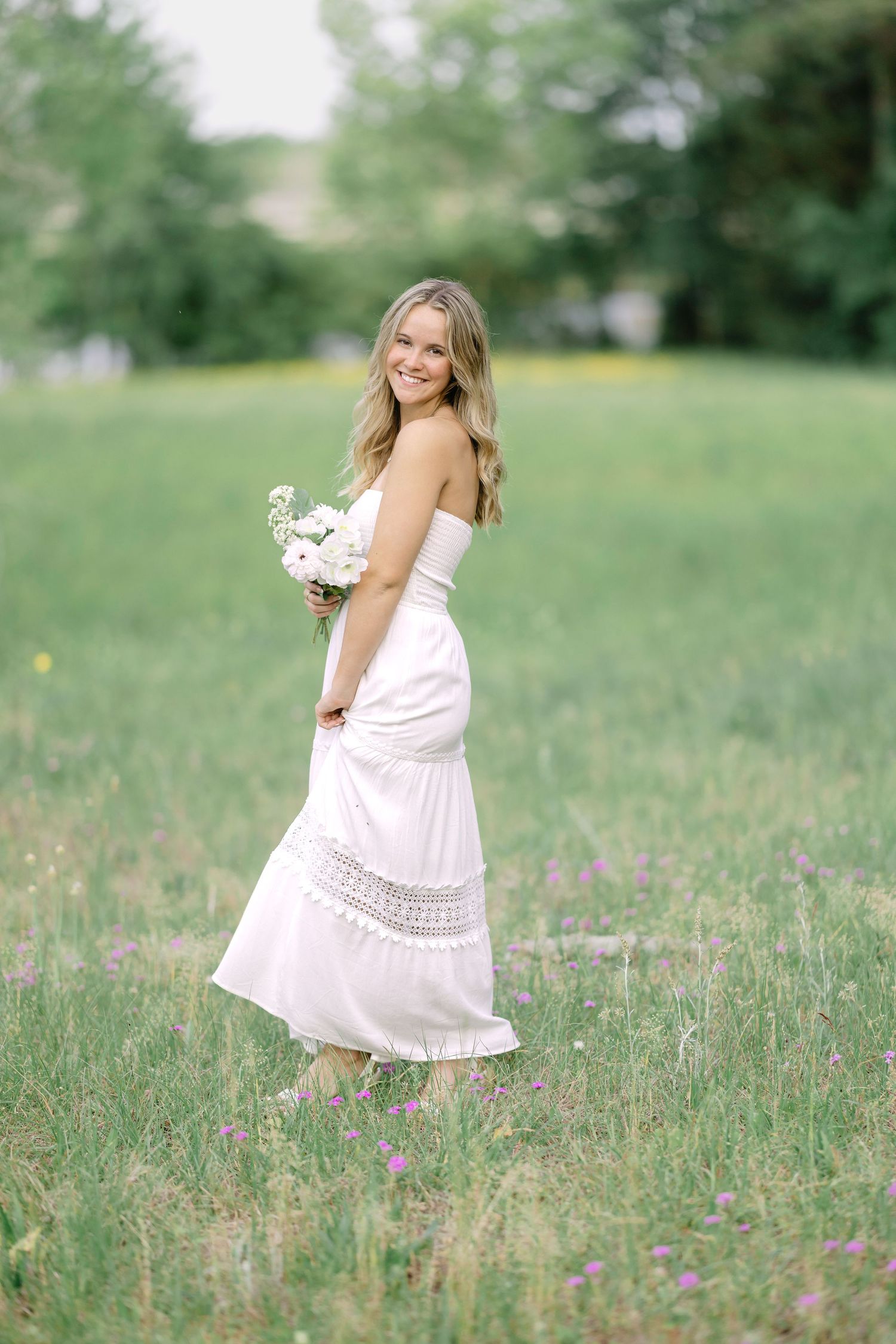 Smiling blonde woman in white strapless dress holding white flowers in a green meadow.