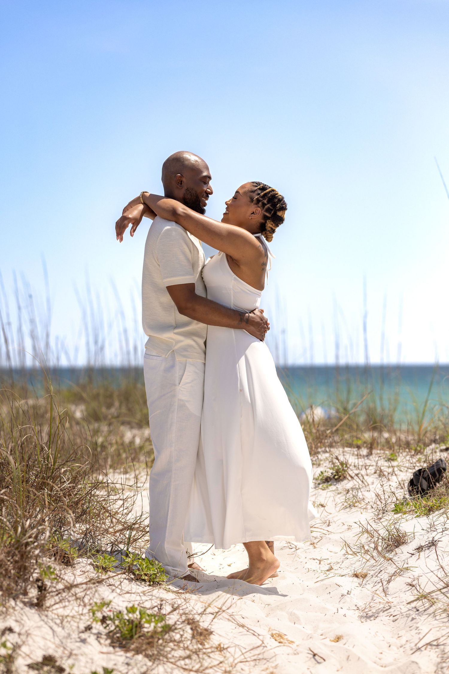A couple in white outfits embracing on a sandy beach with ocean and tall grass in the background on a sunny day.