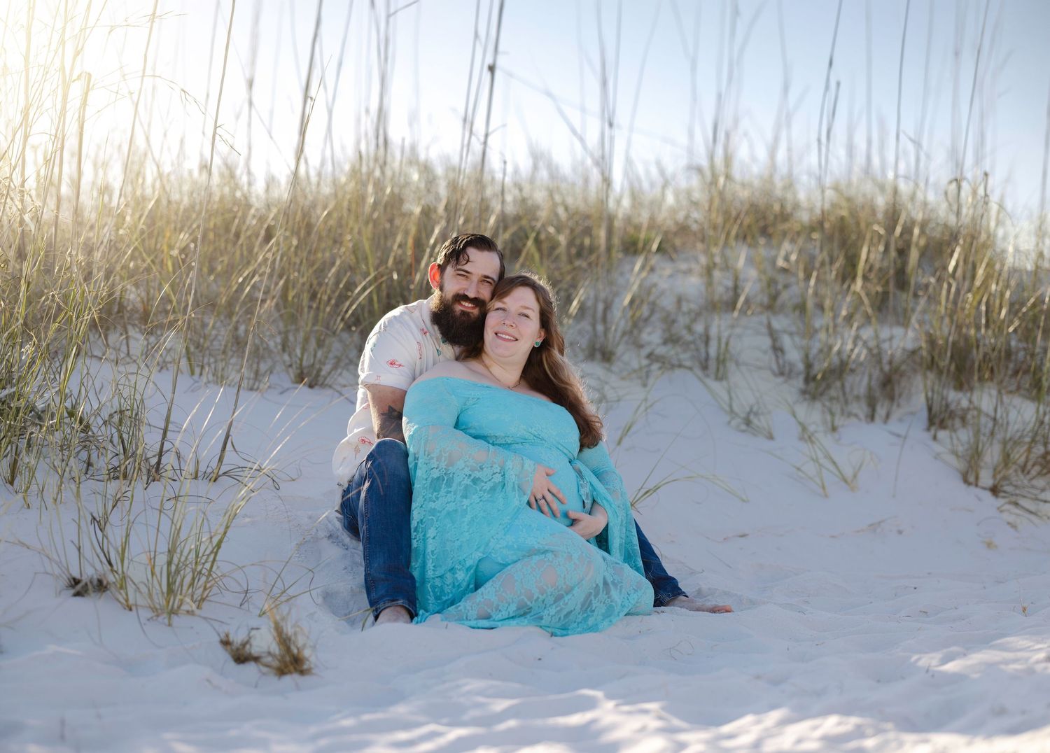 Couple sitting on white sand dunes with sea oats, woman in teal maternity dress, man in white shirt, beach setting.