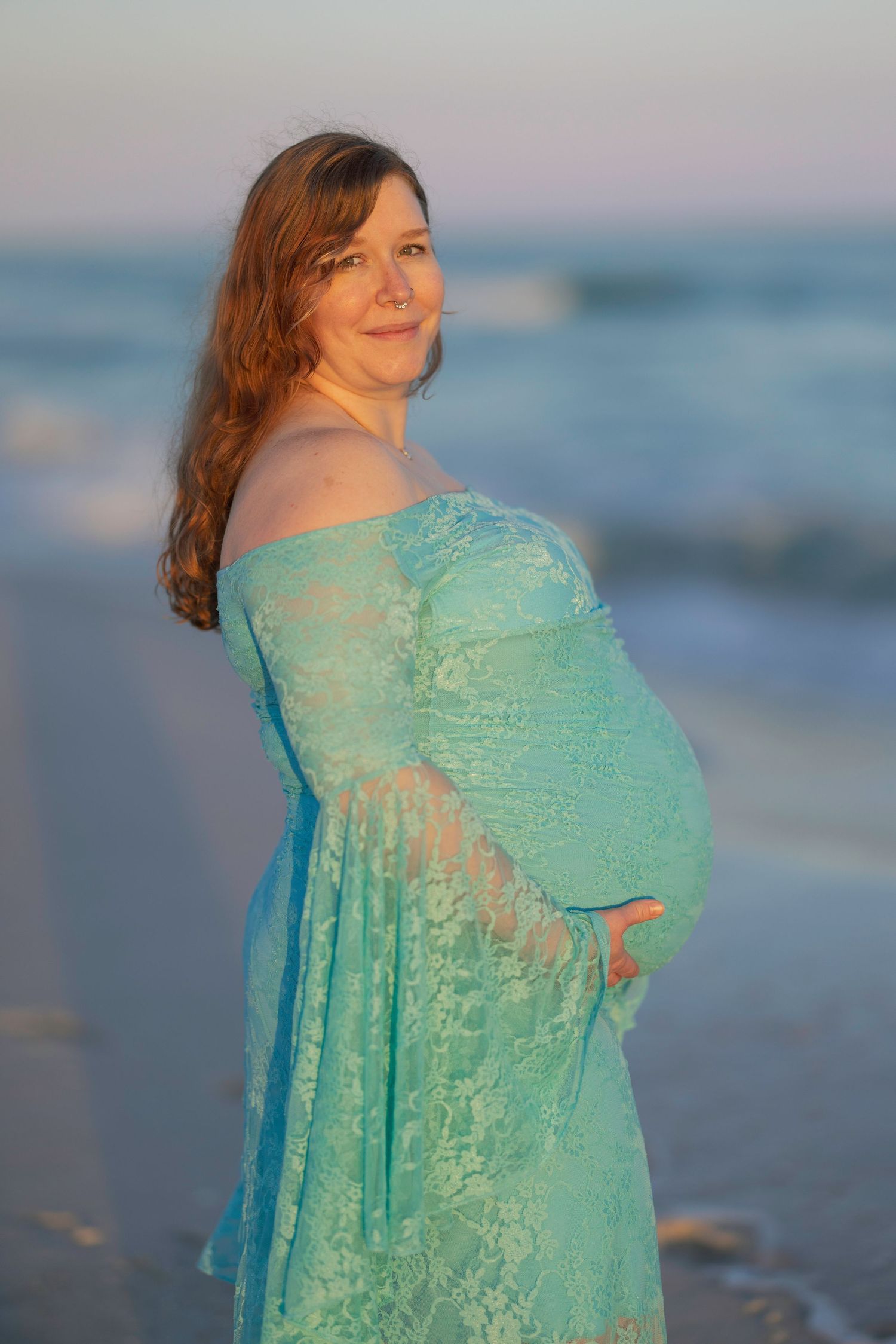 Pregnant woman in teal lace off-shoulder gown poses on beach at sunset, cradling her baby bump.