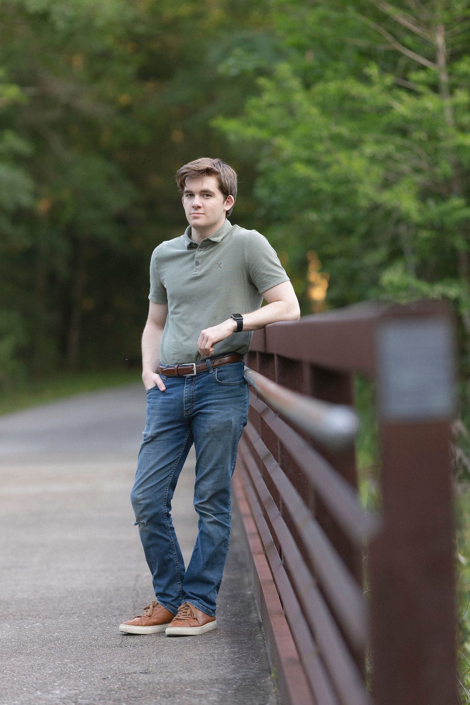 Young man in olive polo and jeans leaning on wooden bridge railing in lush green park setting.