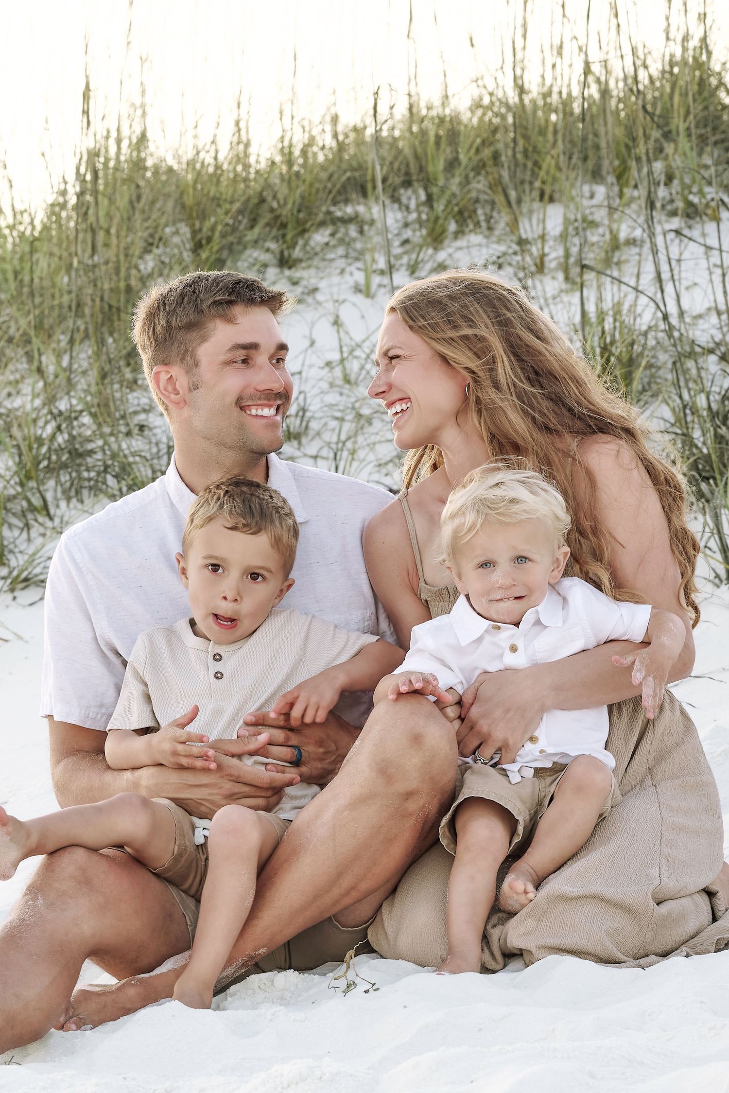 A happy young family of four sits together on white sand dunes at a beach during golden hour.