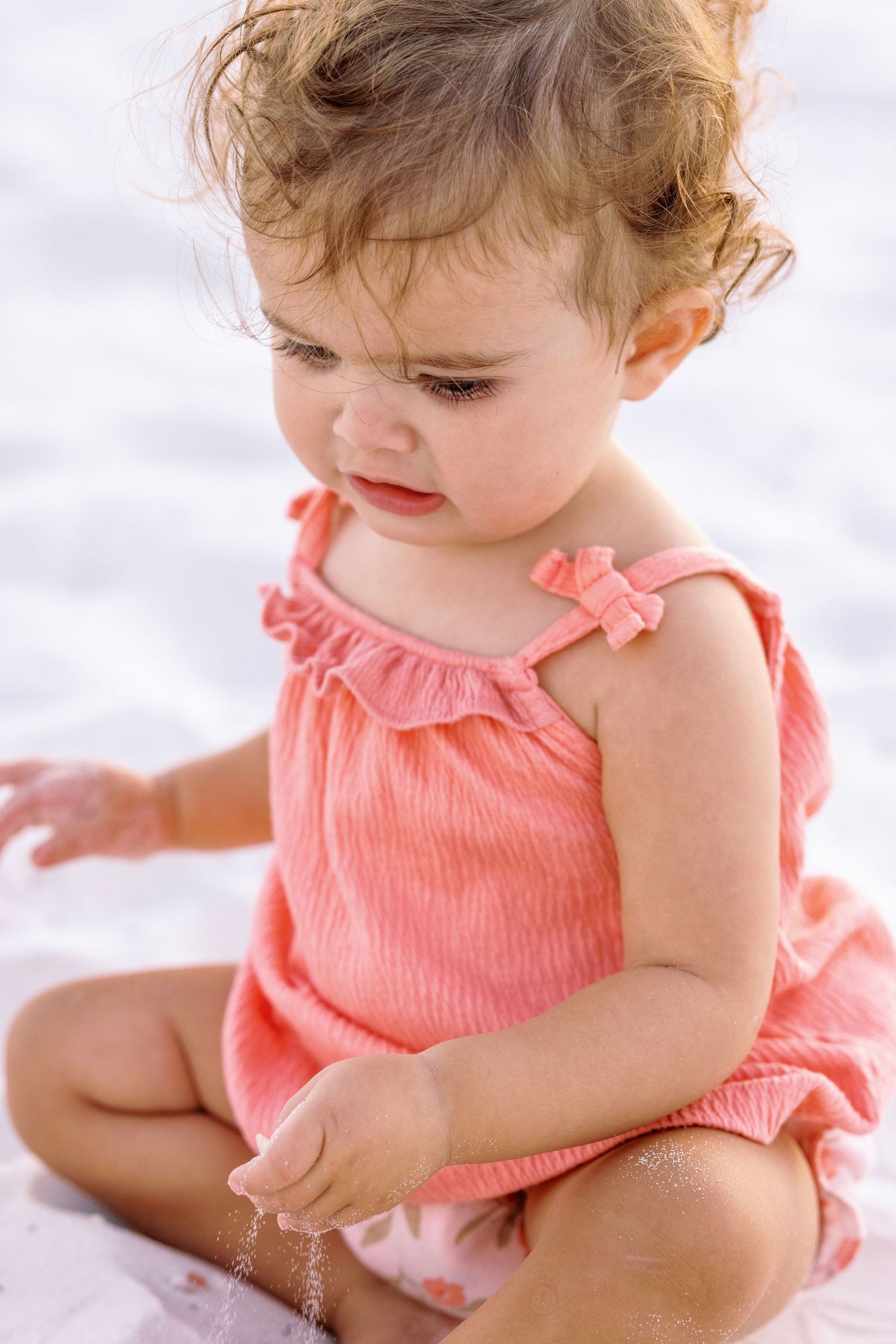 Baby in coral romper sits and plays in soft white sand at the beach.