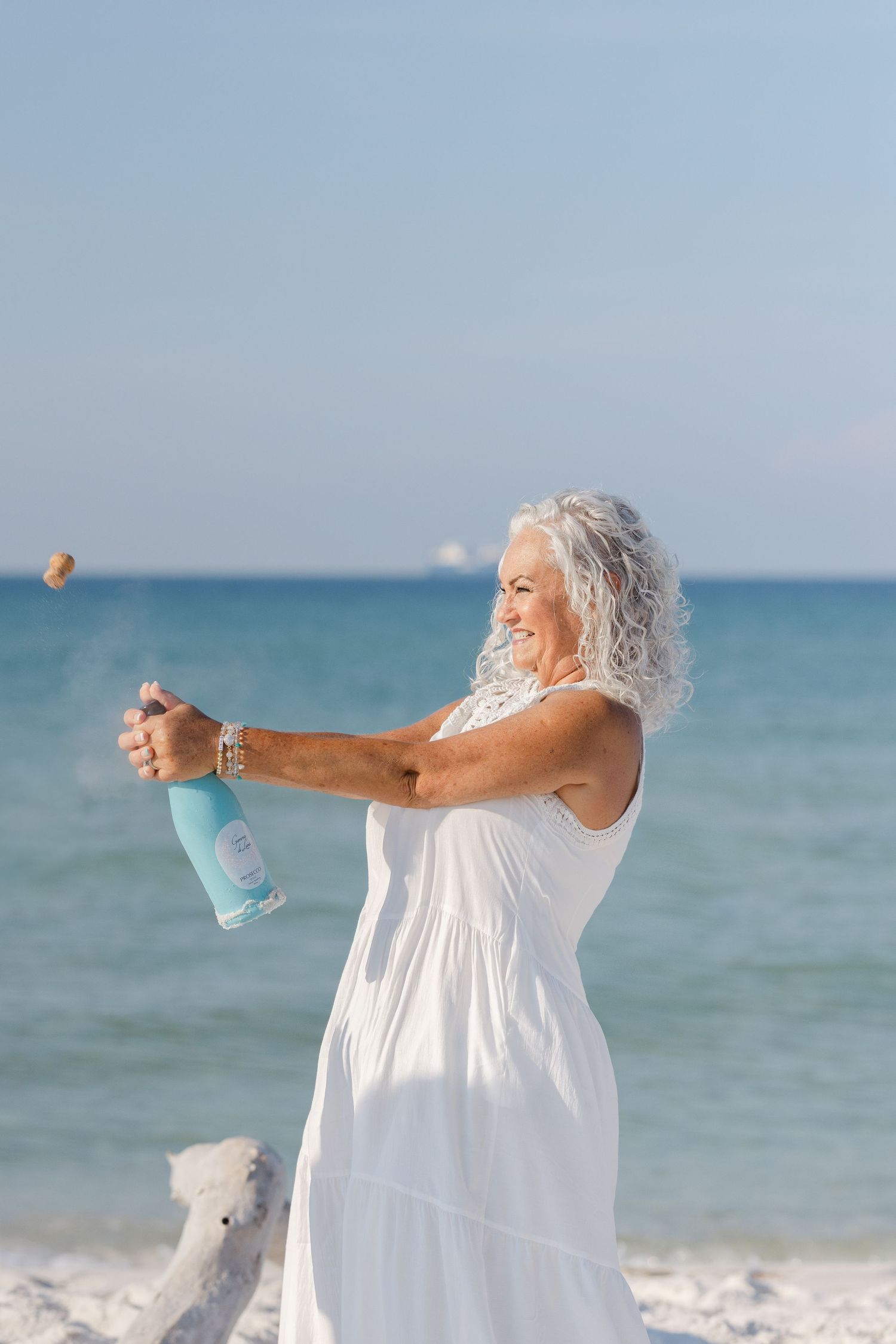 Person in flowing white sundress celebrating on a sunny beach while holding a water bottle.