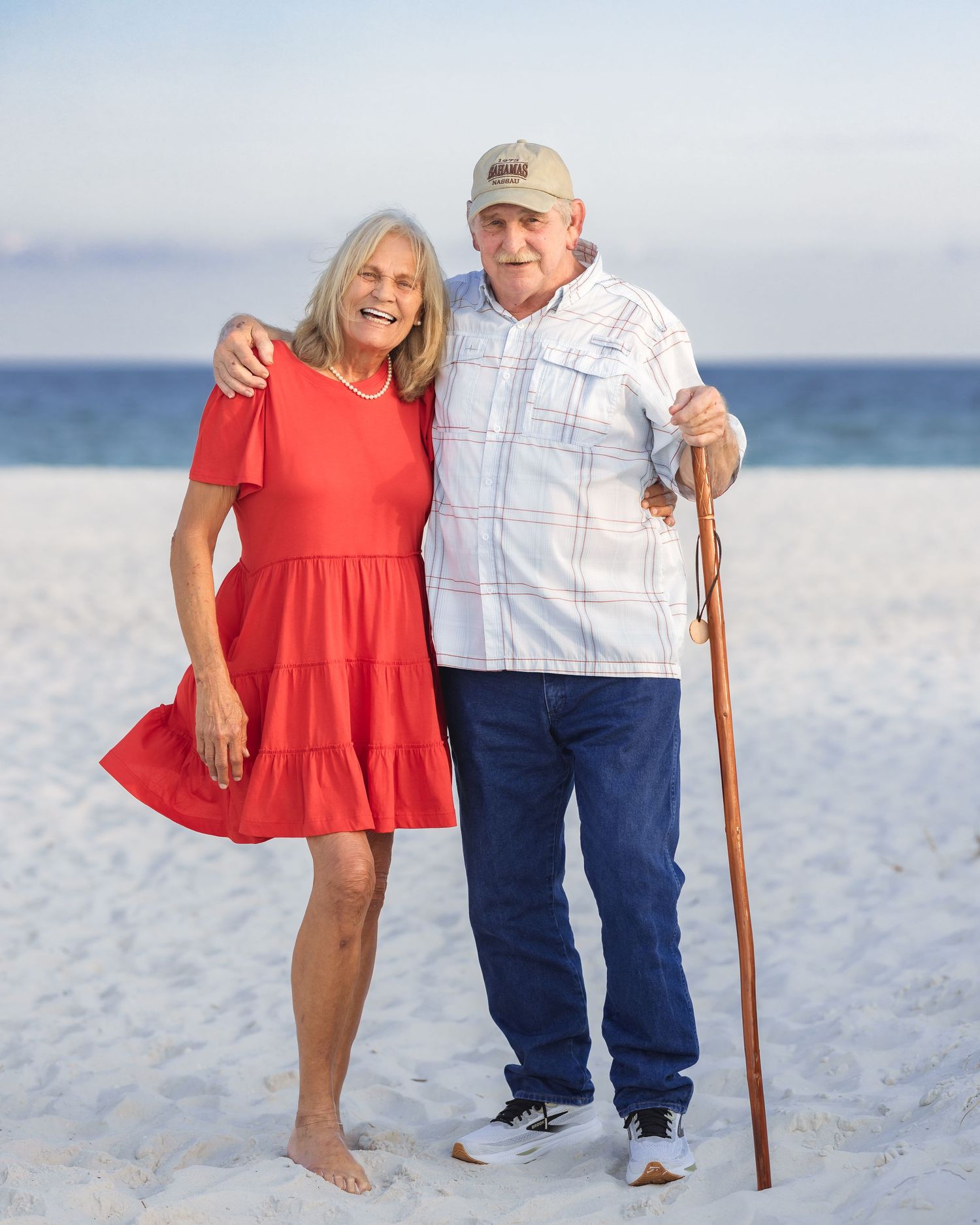 Smiling couple posing on a white sand beach, woman in red dress and man with walking stick, ocean in background.