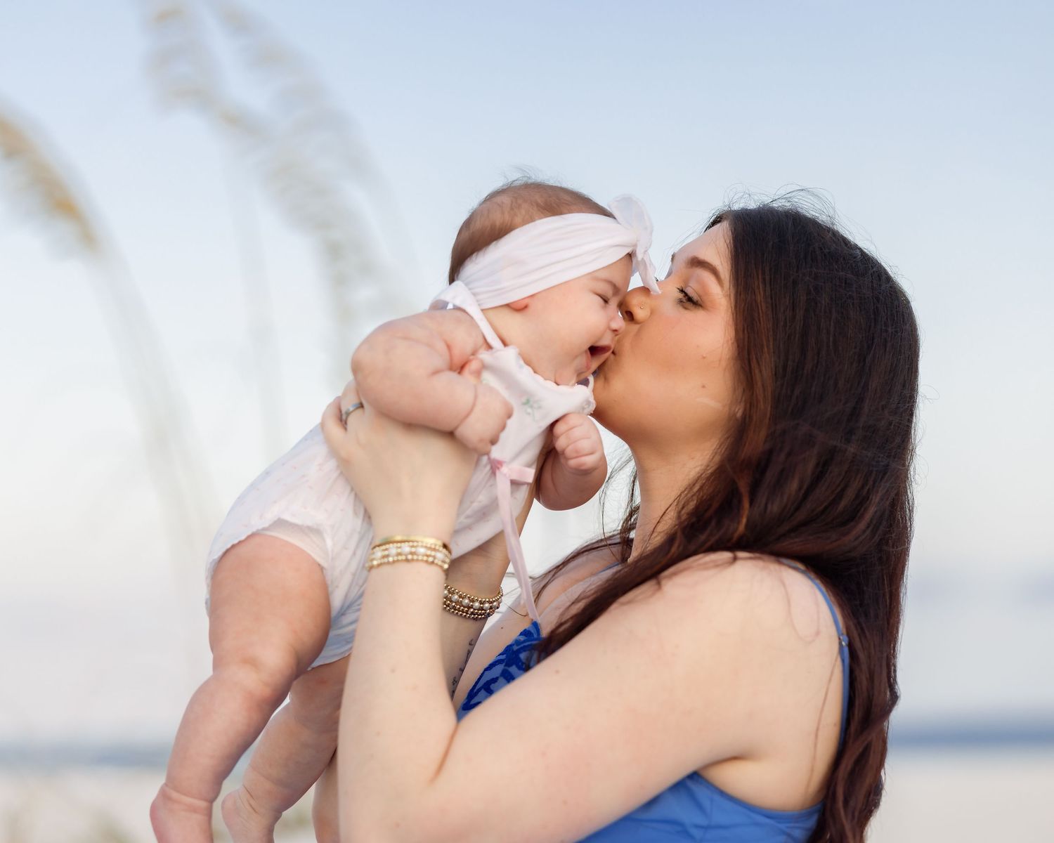 Mother in blue dress kissing her baby girl in white outfit on a sunny beach.