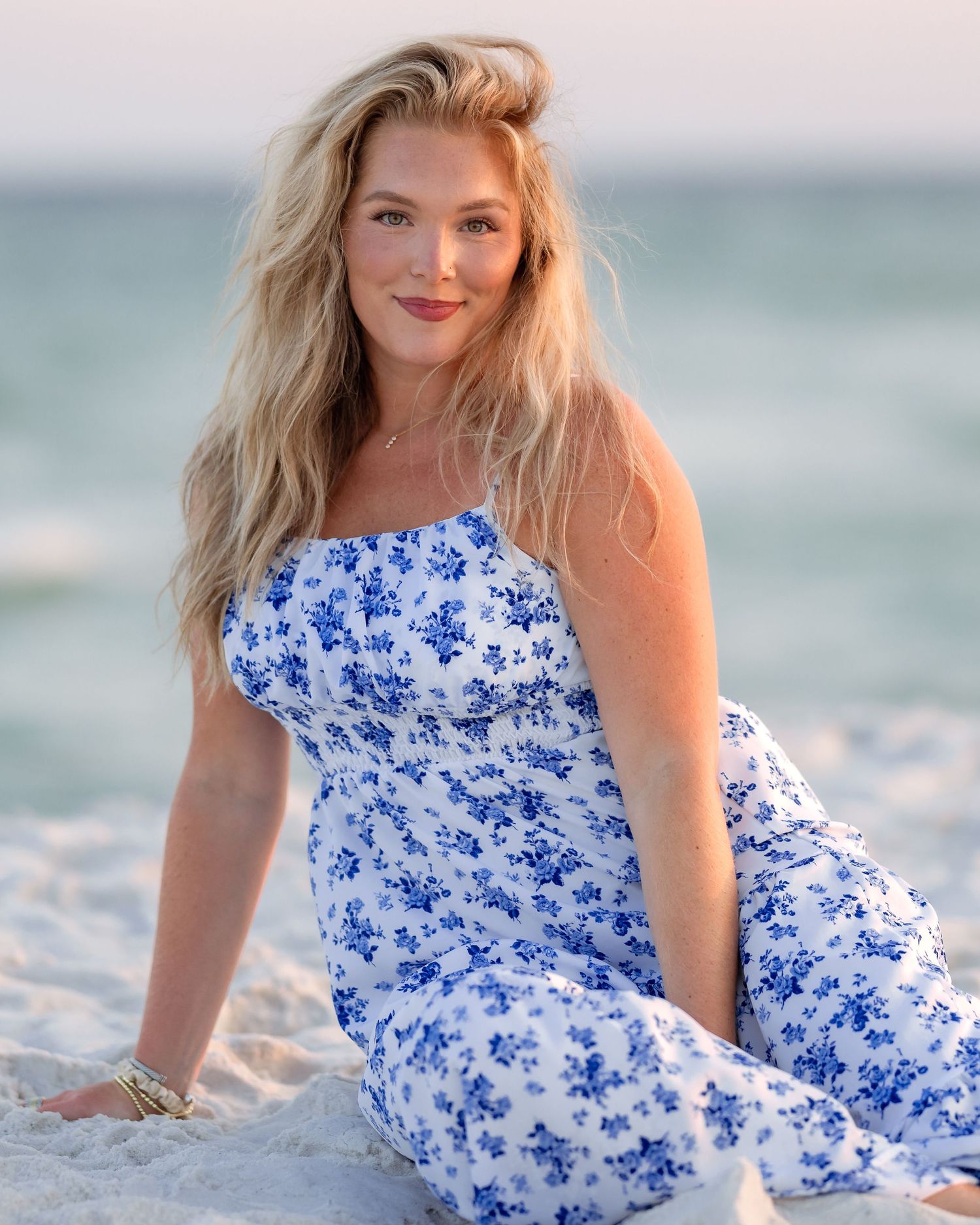 A smiling blonde woman in a blue floral dress sits on white sand beach.