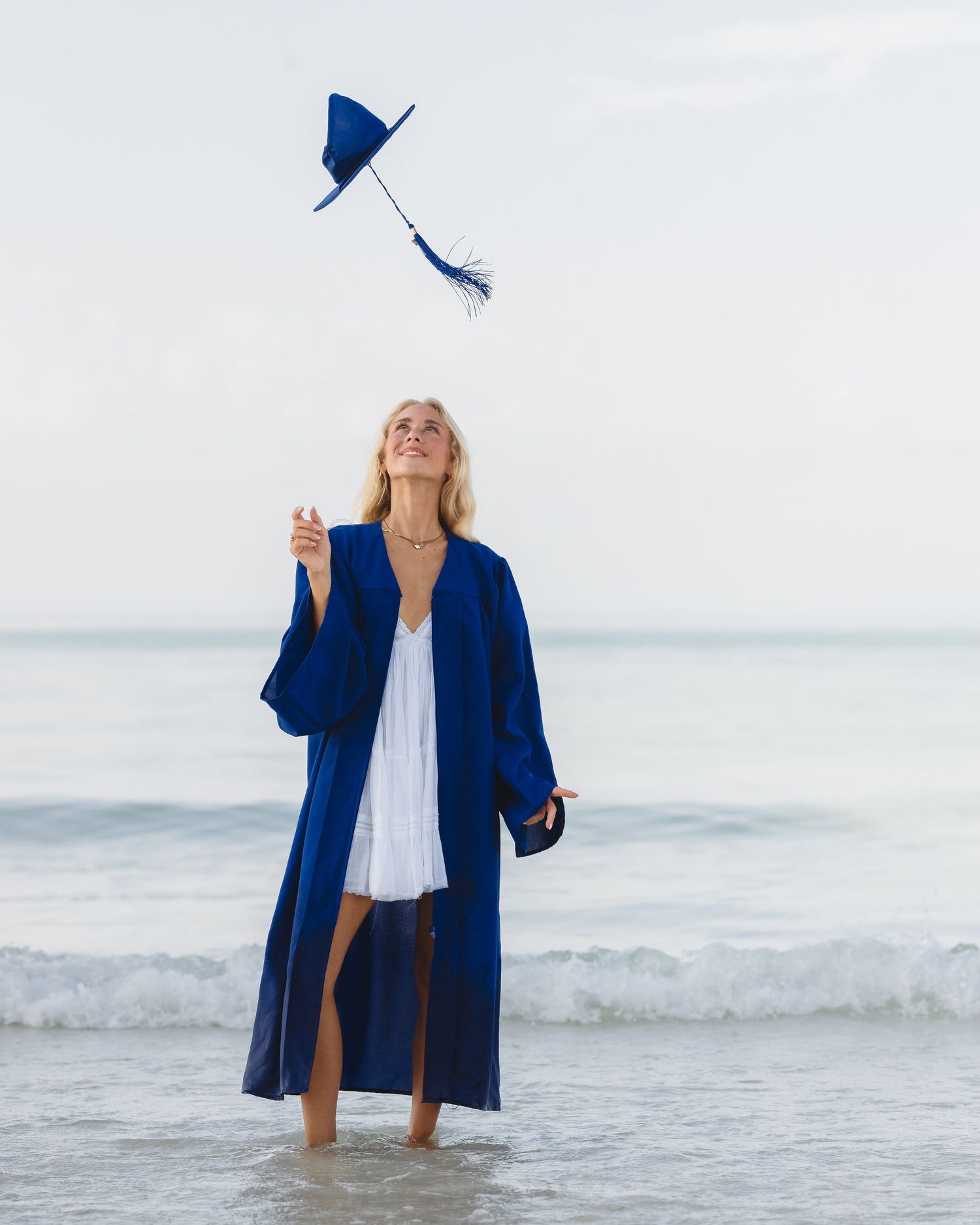 Woman in blue graduation gown tossing cap on beach, wearing white dress, celebrating by the ocean.