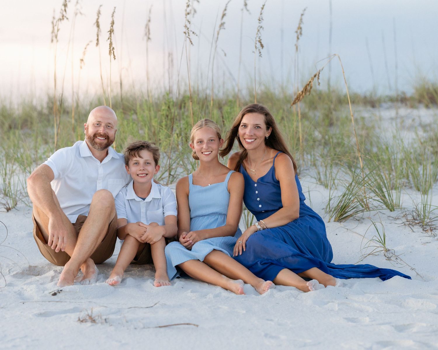 A happy family of four sits together on a sandy beach with sea oats in the background.