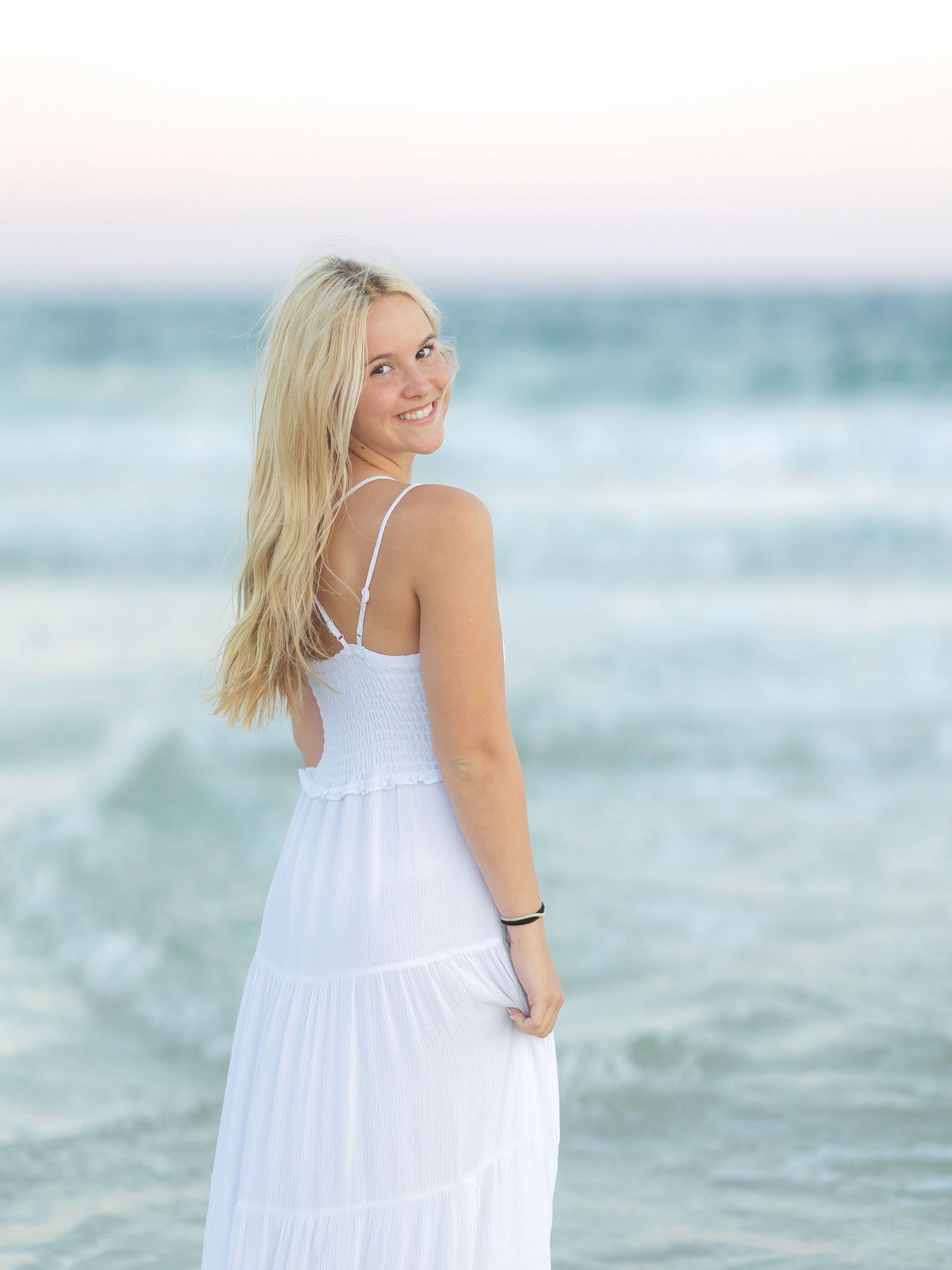 Woman in flowing white maxi dress smiles over shoulder on beach at sunset.