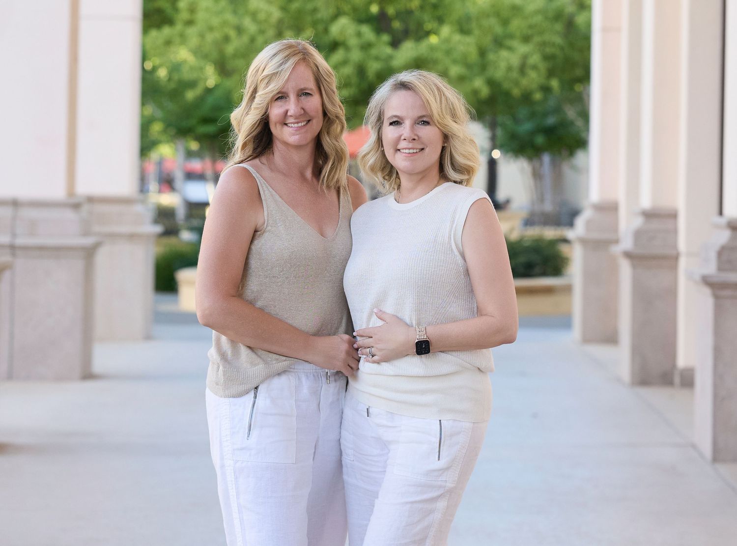 Three women in white dresses stand together smiling outdoors on a sunny day.