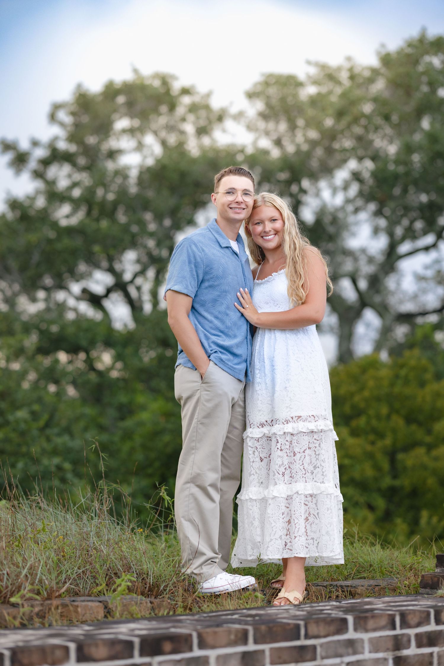 Happy couple embracing outdoors, man in blue polo and woman in white lace maxi dress with green trees behind.