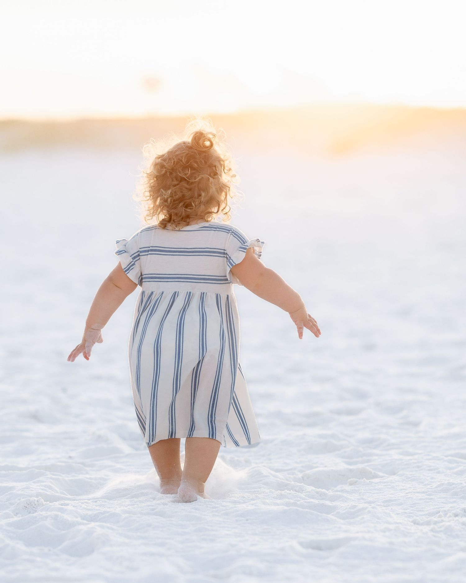 Toddler girl in striped dress walking barefoot on white sand beach at golden sunset.